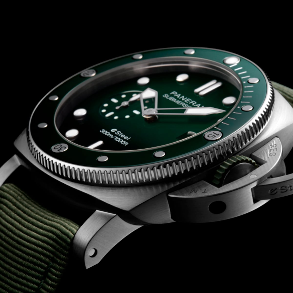 2025 © Panerai Eine Nahaufnahme einer Panerai Submersible Uhr mit grünem Zifferblatt und Lünette, silbernen Indizes und einem strukturierten grünen Armband vor einem schwarzen Hintergrund.