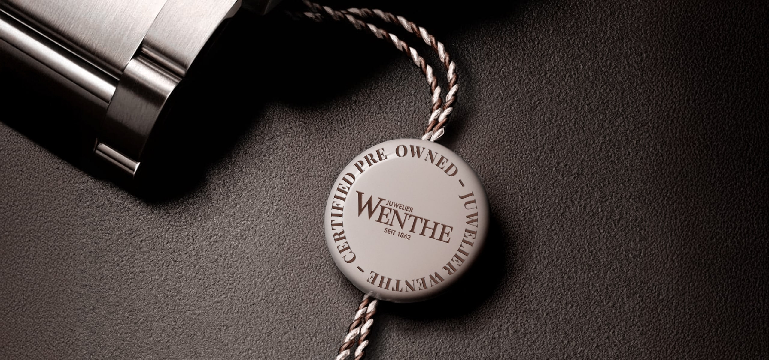 © Juwelier Wenthe Ein rundes weißes Zertifizierungsschild mit dem Aufdruck "Certified Pre-Owned Juwelier Wenthe", das an einer geflochtenen Schnur befestigt ist, liegt auf einer dunklen, strukturierten Oberfläche neben einem glänzenden Metallobjekt.