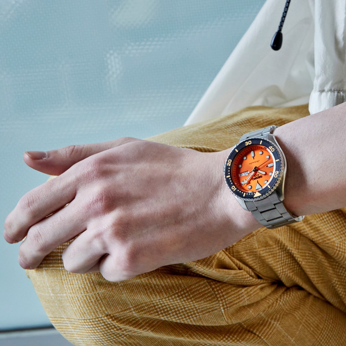 2025 © SEIKO Eine Person, die eine gelb karierte Hose und eine silberne Seiko 5 Sports Armbanduhr mit orangefarbenem Zifferblatt und schwarzer Lünette trägt, stützt ihre Hand auf ihr Knie vor einem unscharfen Hintergrund.