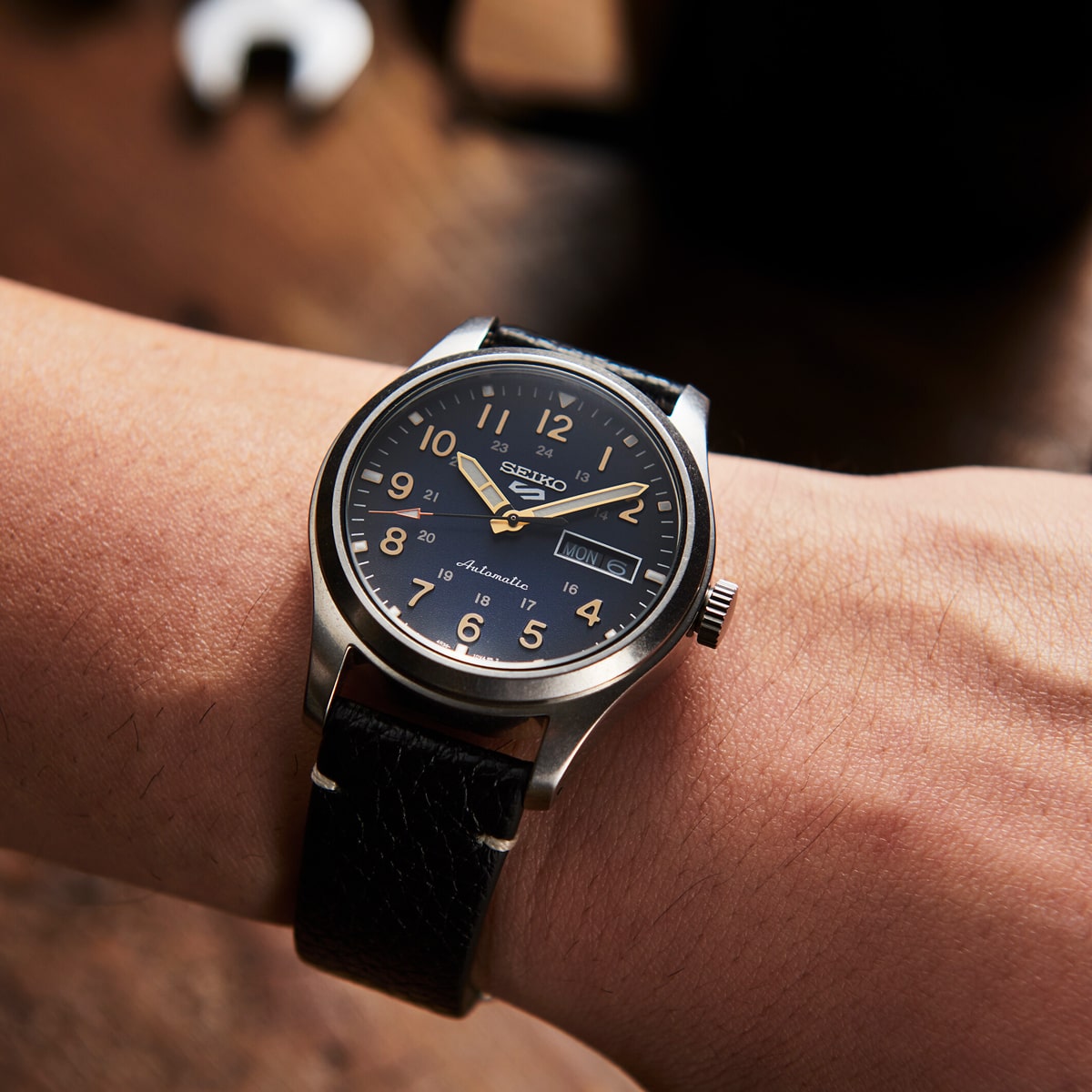 2025 © SEIKO Eine Nahaufnahme des Handgelenks einer Person, die eine Seiko 5 Sports Automatikuhr mit schwarzem Zifferblatt, silbernem Gehäuse, schwarzem Lederarmband und arabischen Ziffern trägt. Der Hintergrund ist unscharf mit einer warmen, hölzernen Oberfläche.