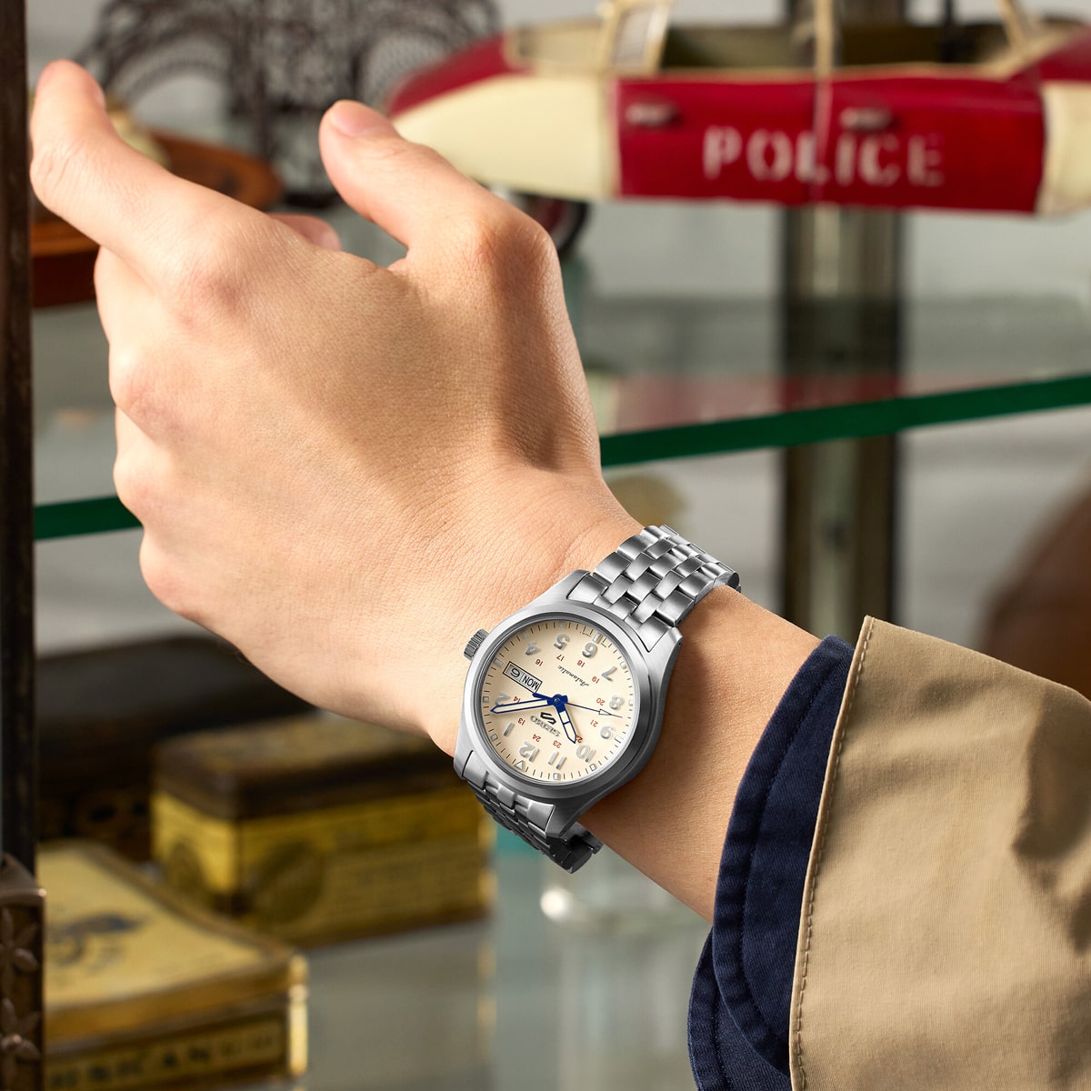 2025 © SEIKO Eine Person, die eine silberne Seiko 5 Sports Armbanduhr mit cremefarbenem Ziffernblatt trägt, streckt ihren linken Arm vor einem Glasregal aus, in dem alte Gegenstände ausgestellt sind, darunter ein rot-weißes Polizeimodell.