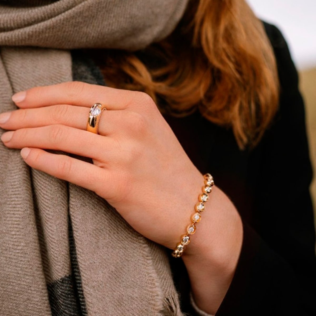 2025 © SCHAFFRATH Nahaufnahme einer Frauenhand, die einen Goldring mit einem klaren Stein und einen SCHAFFRATH-Armschmuck - ein Goldarmband mit runden Edelsteinen - trägt. Sie ist mit einem dunklen Mantel und einem beigen Schal bekleidet, ihr Haar ist teilweise sichtbar.
