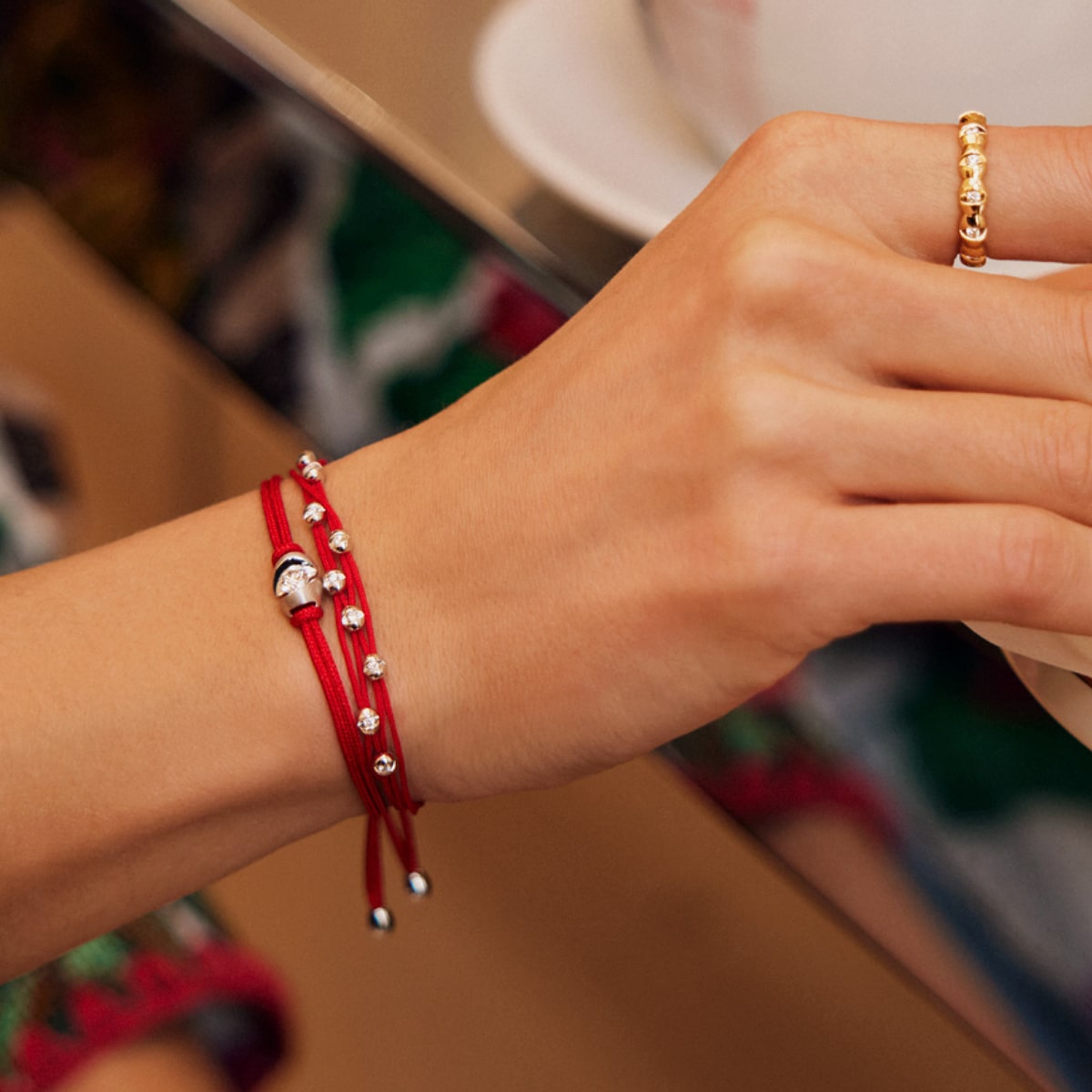 2025 © SCHAFFRATH Eine mit einem goldenen Perlenring und SCHAFFRATH-Armschmuck - einem roten Schnurarmband mit kleinen Silberperlen - geschmückte Hand ruht auf einer spiegelnden Fläche neben einer weißen Tasse.
