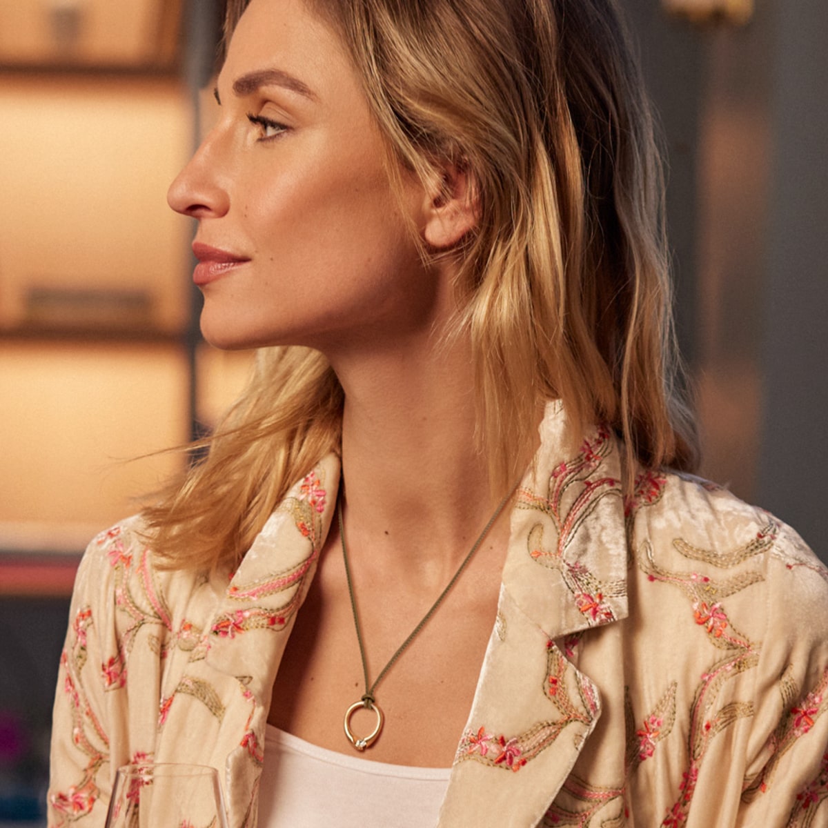 2025 © SCHAFFRATH Eine Frau mit blondem Haar, die einen hellen, mit Blumen bestickten Blazer und eine runde Halskette mit Anhänger von SCHAFFRATH Halsschmuck trägt, blickt in einem sanft beleuchteten Innenraum zur Seite.