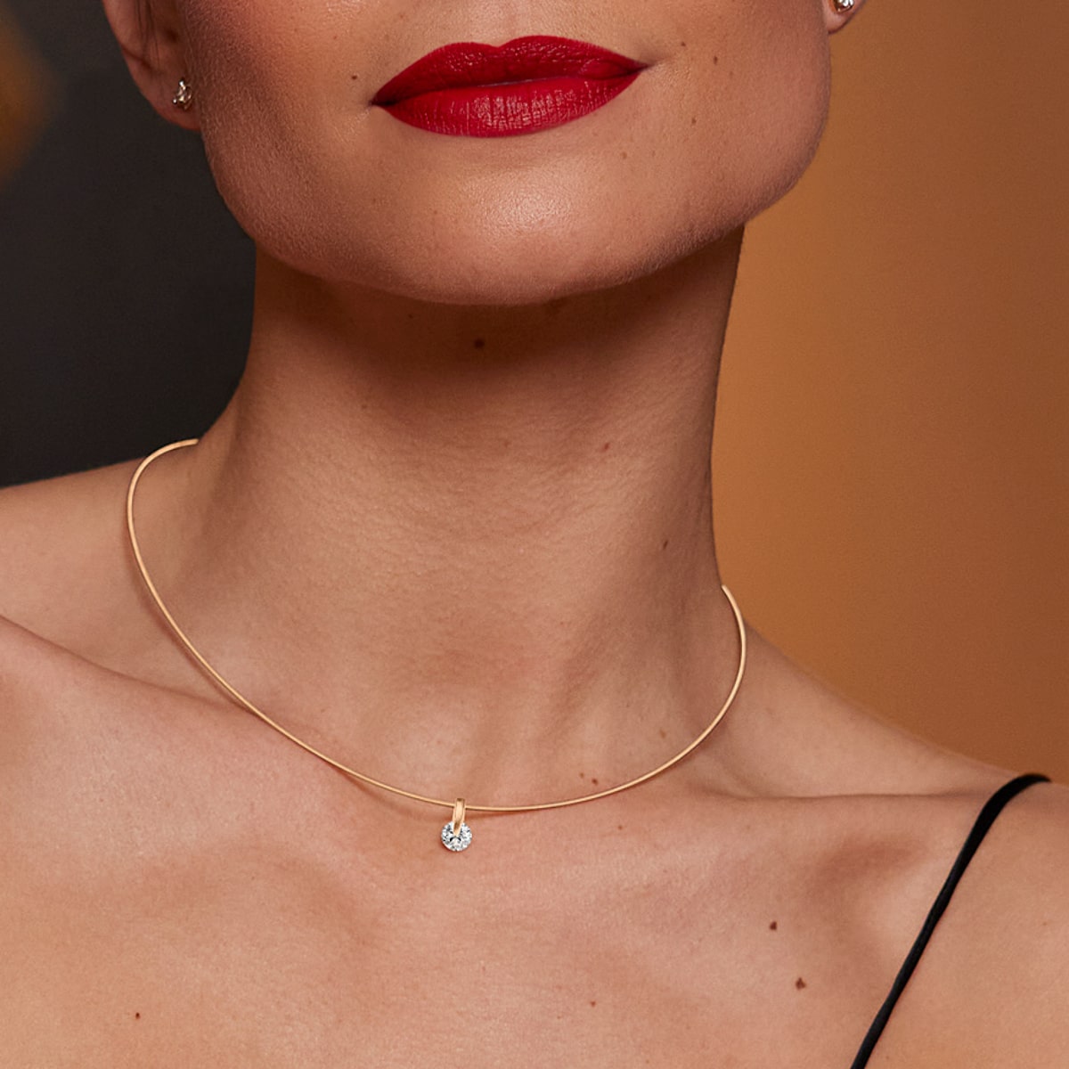 2025 © SCHAFFRATH Eine Frau mit leuchtend rotem Lippenstift und einer dünnen goldenen Halskette mit einem einzelnen runden Diamantanhänger von SCHAFFRATH Halsschmuck, von den Schultern aufwärts vor einem sanft verschwommenen Hintergrund gezeigt.