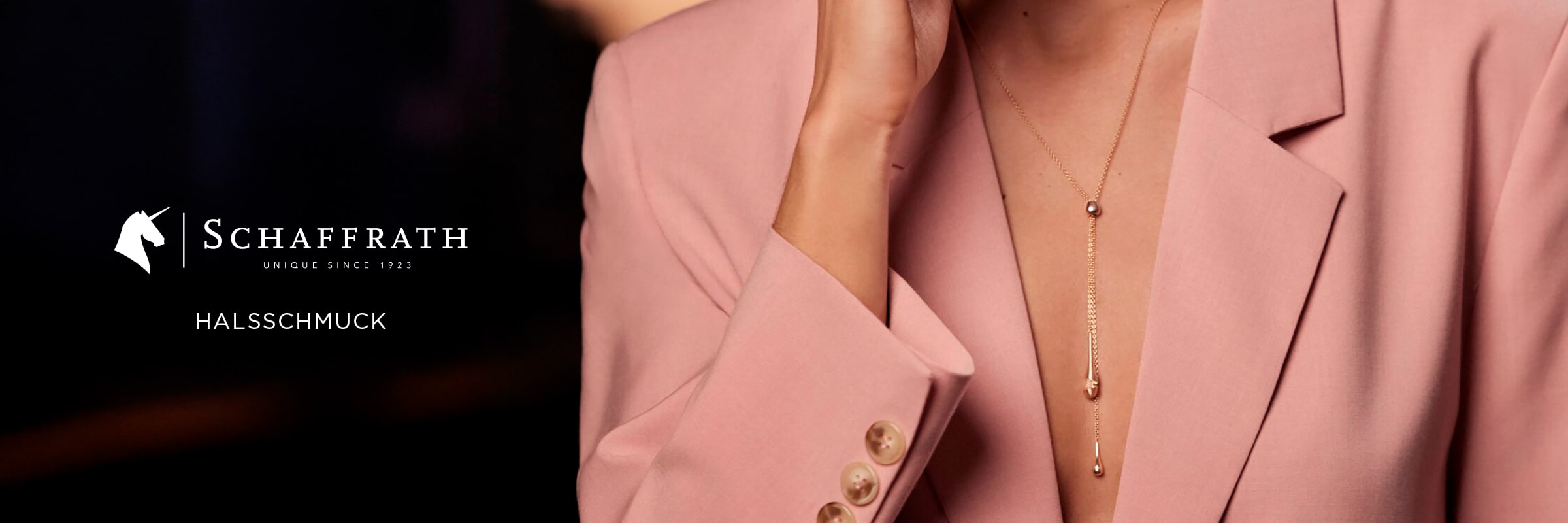2025 © SCHAFFRATH Eine Frau in einem rosafarbenen Blazer trägt zwei zarte SCHAFFRATH Halsschmuck Goldketten mit Anhängern. Der Hintergrund ist unscharf, mit dem Schaffrath-Logo und dem Wort "Halsschmuck" auf der linken Seite des Bildes.