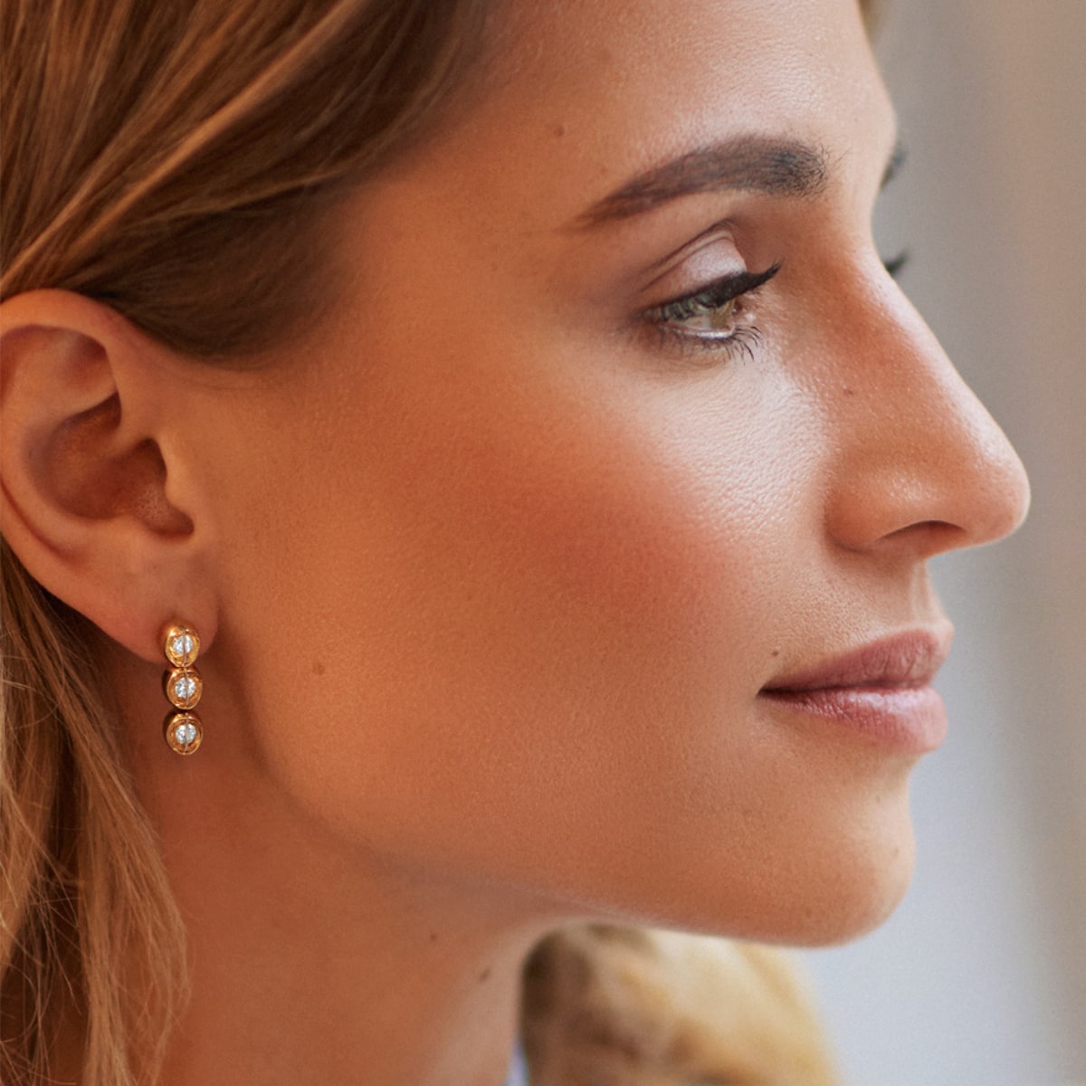 2025 © SCHAFFRATH Eine Frau mit heller Haut und blondem Haar ist im Profil abgebildet. Sie trägt elegante SCHAFFRATH Ohrringe aus Gold mit drei runden Edelsteinen. Sie hat einen ruhigen Ausdruck und ein natürliches Make-up.