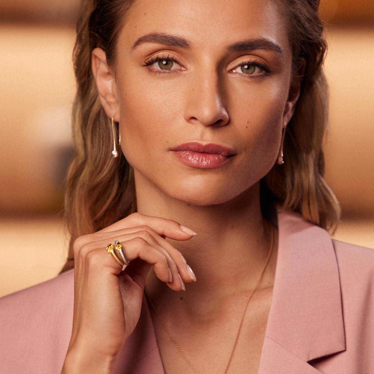 2025 © SCHAFFRATH Eine Frau mit hellbraunem Haar und haselnussbraunen Augen trägt einen rosafarbenen Blazer, Goldschmuck, SCHAFFRATH Ringe und Perlenohrringe und blickt selbstbewusst mit neutralem Blick in die Kamera.