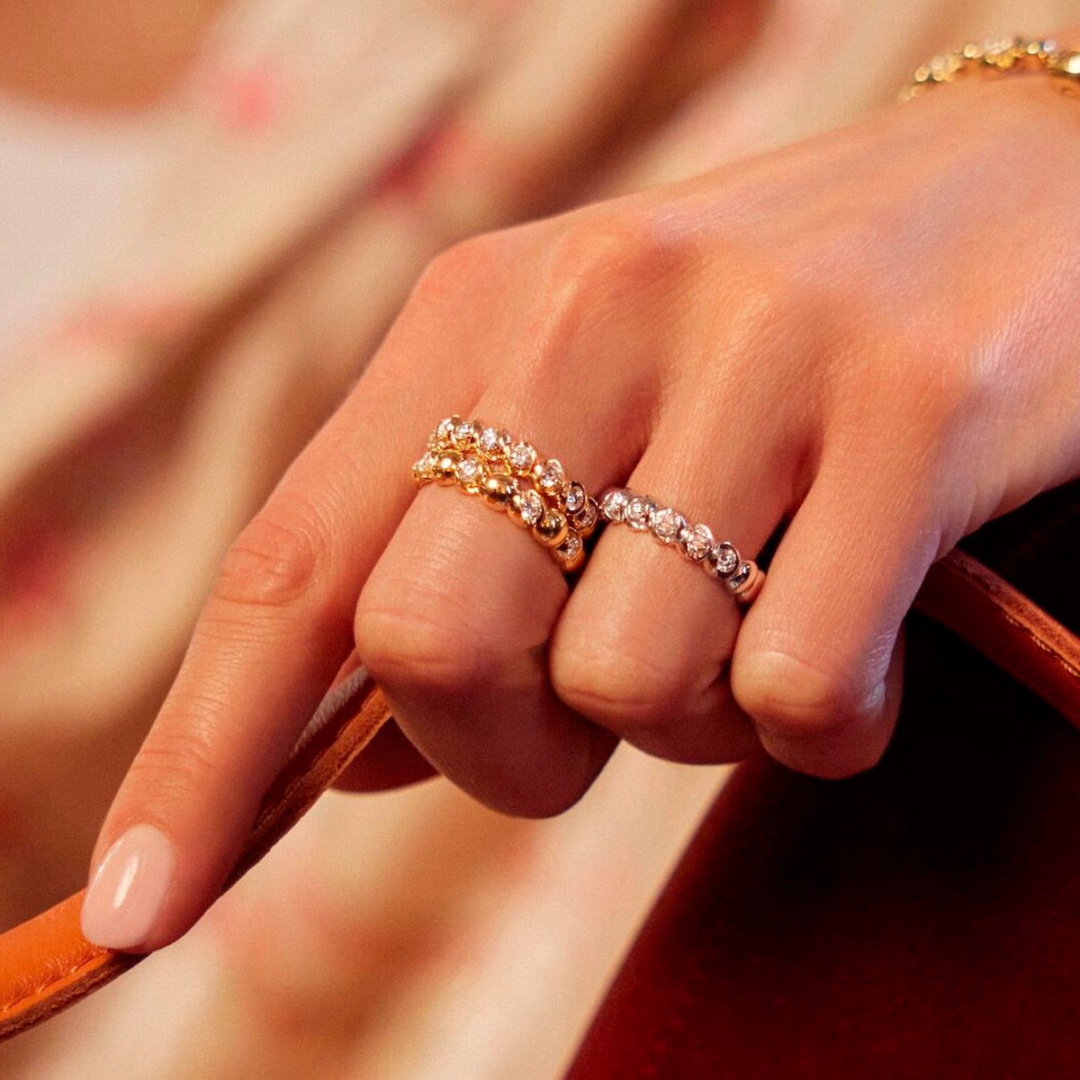 2025 © SCHAFFRATH Nahaufnahme einer Hand, die auf einem braunen Armband ruht und zwei SCHAFFRATH Ringe mit funkelnden klaren Steinen am Zeige- und Mittelfinger trägt. Die Nägel sind mit einem blassrosa Lack ordentlich manikürt.