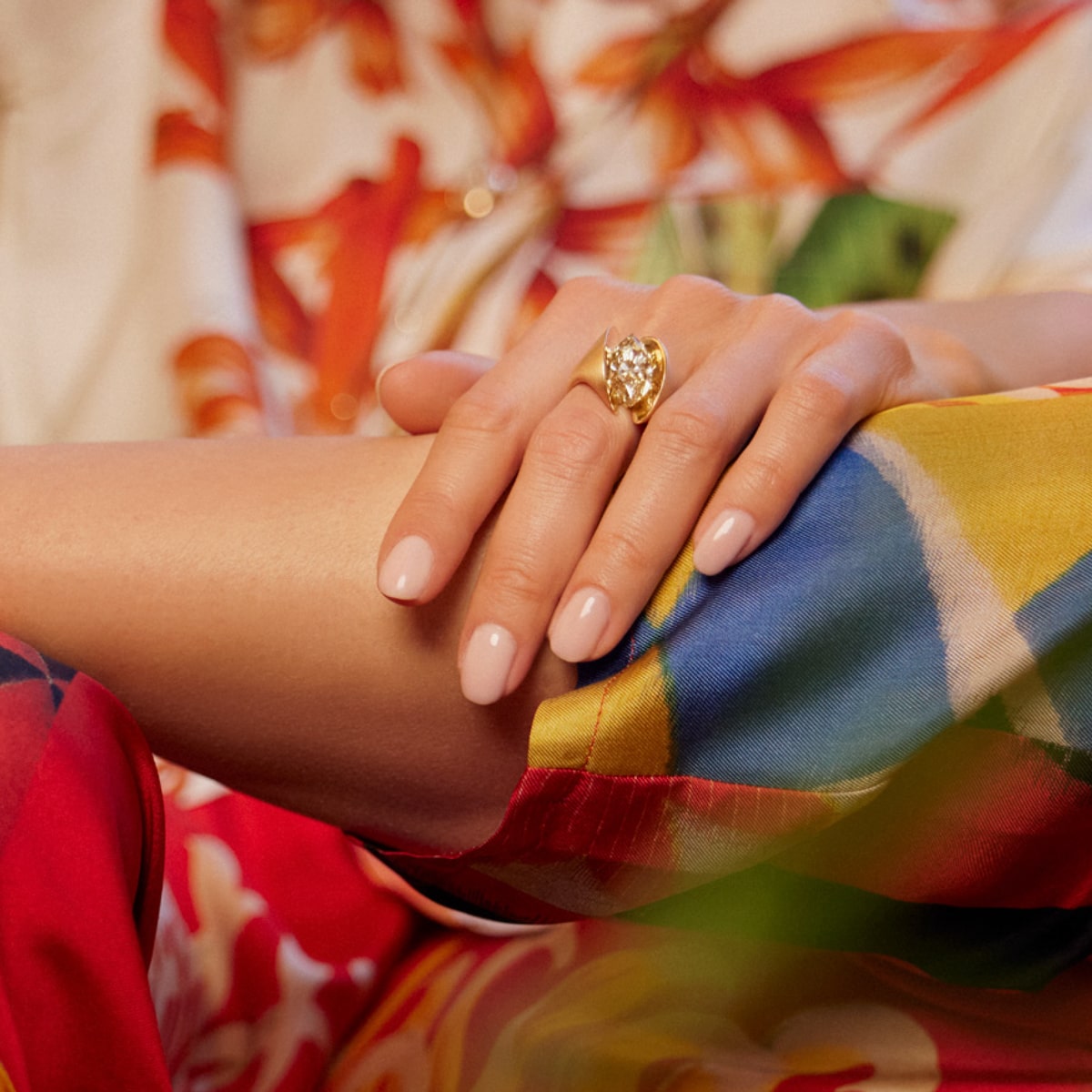2025 © SCHAFFRATH Nahaufnahme einer Frauenhand mit sauber manikürten, nackten Nägeln, die mit einem auffälligen SCHAFFRATH Ringe-Goldring verziert ist, der sanft auf ihrer bunten, blumengemusterten Kleidung ruht.