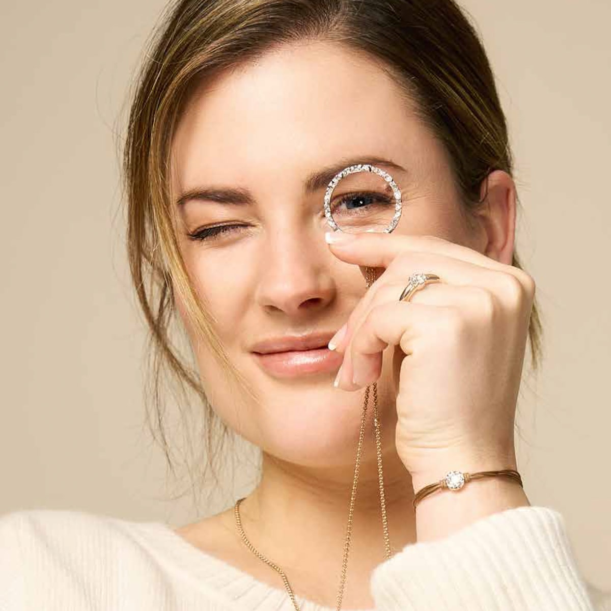 2025 © Schmuckwerk Eine Frau mit hellem Haar hält spielerisch einen Schmuckwerk Halsschmuck-Anhänger an ihr rechtes Auge und blinzelt. Sie trägt ein weißes Oberteil, ein Armband und Ringe vor einem neutralen beigen Hintergrund.