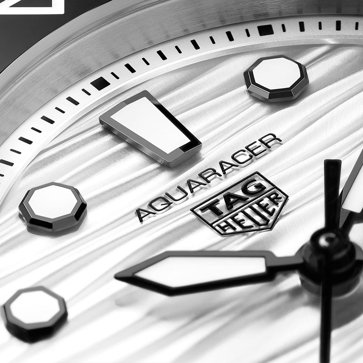 2025 © TAG HEUER Nahaufnahme des Ziffernblatts einer TAG Heuer Aquaracer mit markanten Stundenmarkierungen, ausgeprägten Minuten- und Stundenzeigern und dem Markenlogo auf einem strukturierten silbernen Hintergrund.