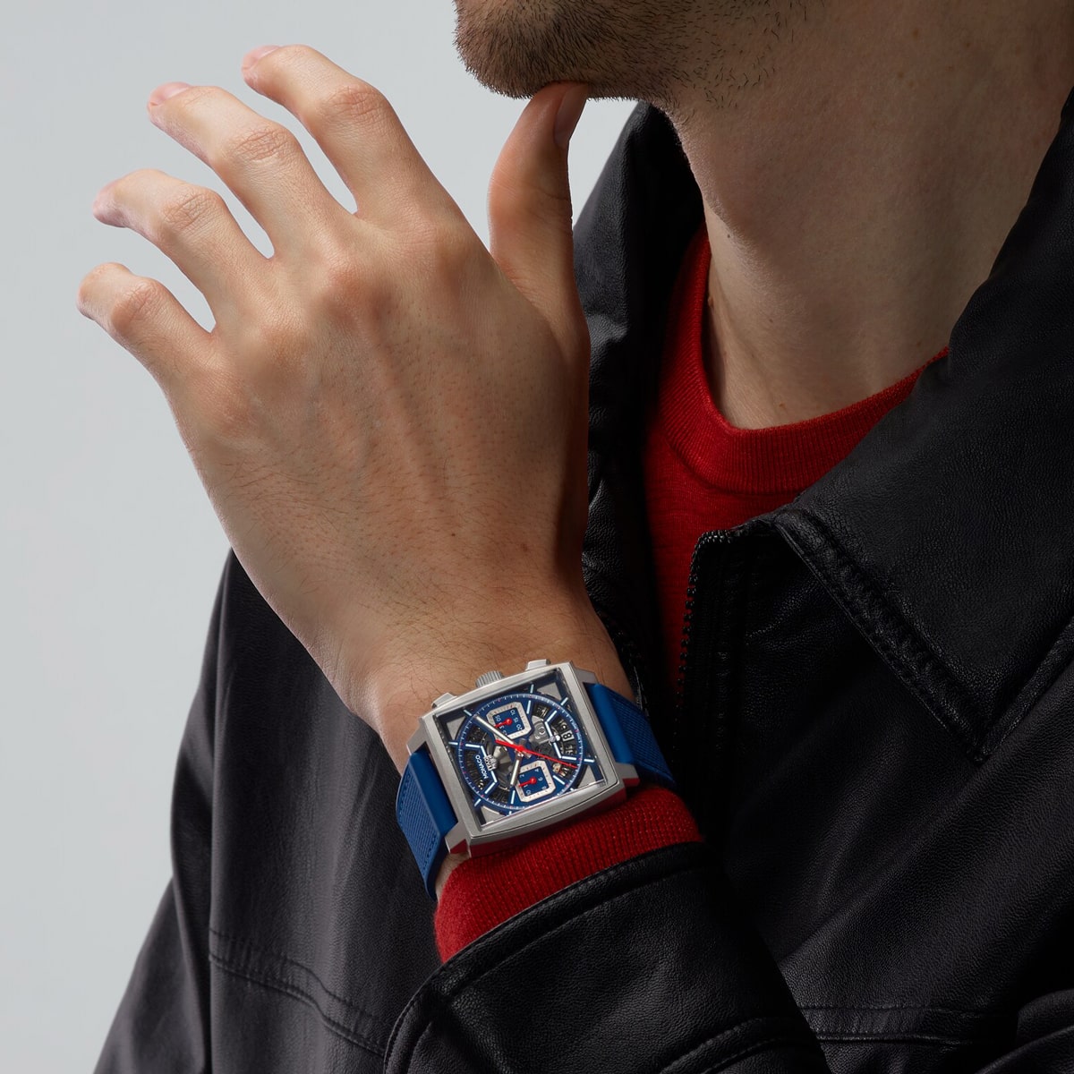 2025 © TAG HEUER Eine Person, die eine schwarze Lederjacke und ein rotes Hemd trägt, berührt ihr Kinn und zeigt eine TAG Heuer Monaco mit einem quadratischen Zifferblatt, einem blauen Armband und einem detaillierten blauen, roten und silbernen Zifferblatt an ihrem Handgelenk.
