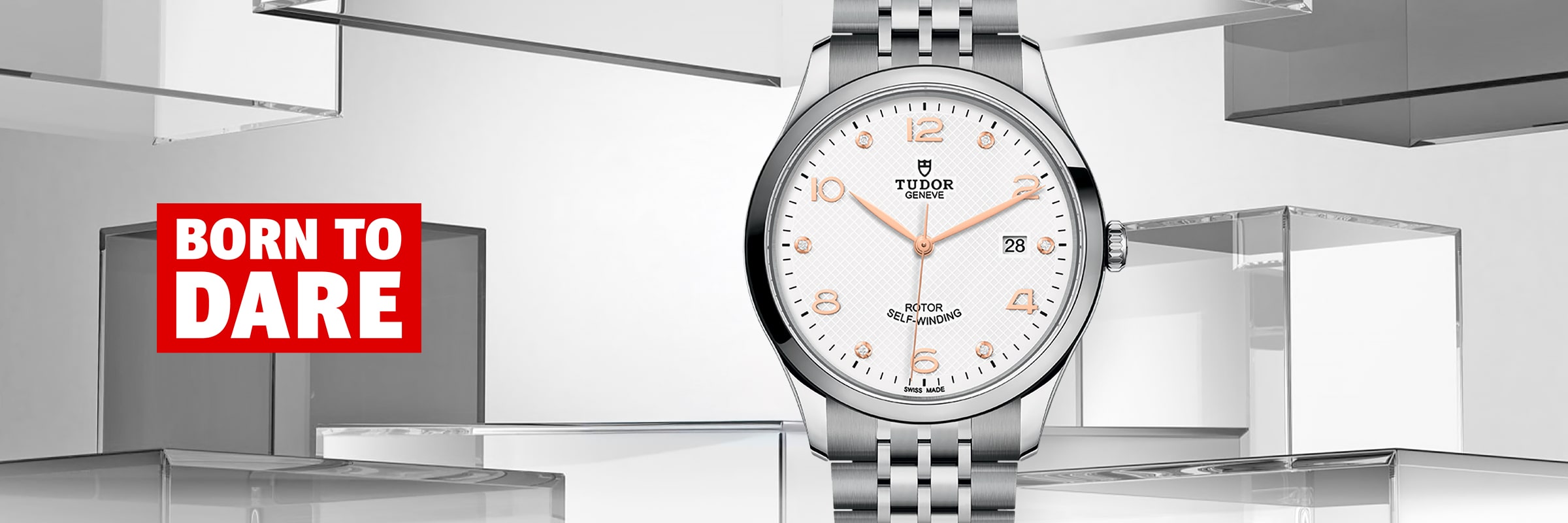 2025 © TUDOR Eine silberne Tudor 1926 Armbanduhr mit weißem Zifferblatt ist zwischen transparenten Blöcken ausgestellt. Die Uhr zeigt die Uhrzeit 10:08 an, und links davon steht in einem roten Kasten "BORN TO DARE" in fetter weißer und roter Schrift.
