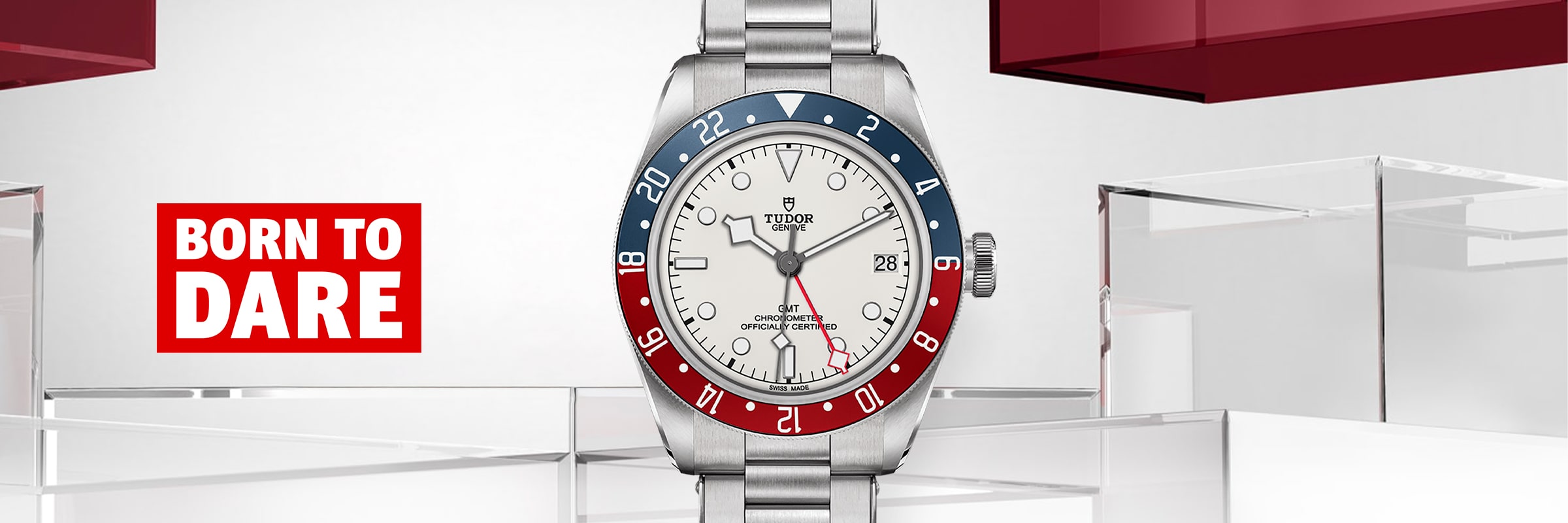 2025 © TUDOR Eine silberne Tudor Black Bay GMT-Armbanduhr mit roter und blauer Lünette und weißem Zifferblatt ist vor einem modernen, geometrischen Hintergrund abgebildet. Die Worte "BORN TO DARE" erscheinen in fetter roter und weißer Schrift auf der linken Seite.