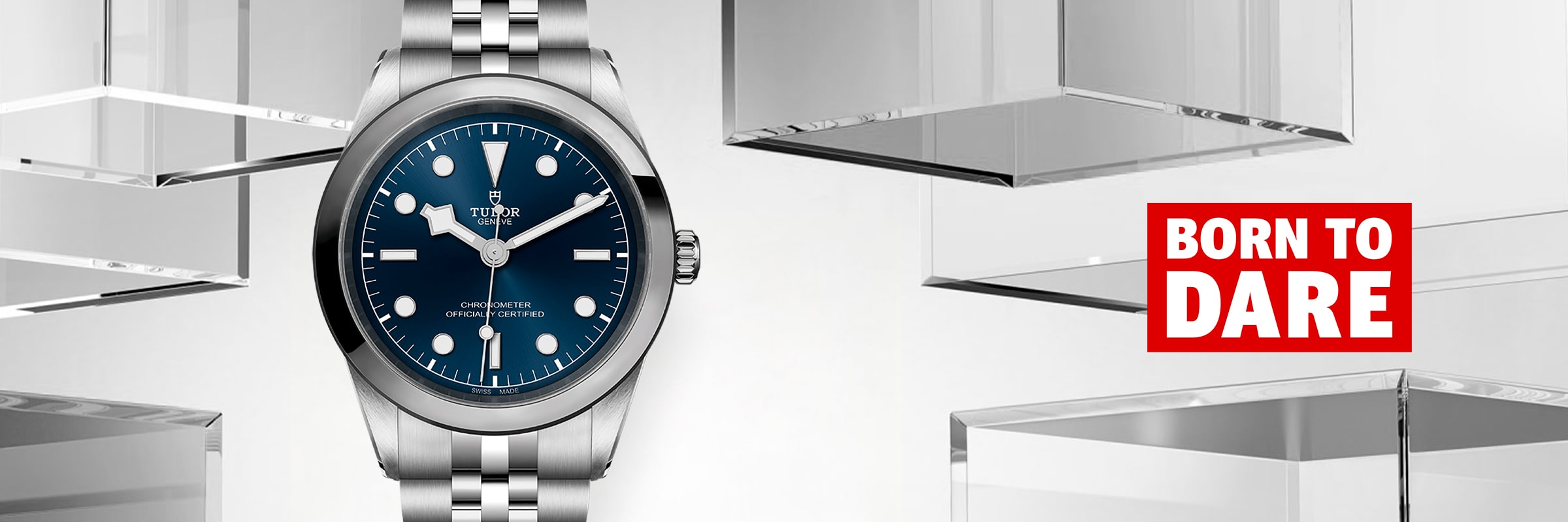 2025 © TUDOR Eine silberne Tudor Black Bay One Armbanduhr mit blauem Zifferblatt ist neben glasähnlichen Würfeln ausgestellt. Neben der Uhr steht in fetten roten und weißen Lettern "BORN TO DARE" auf hellgrauem Hintergrund.
