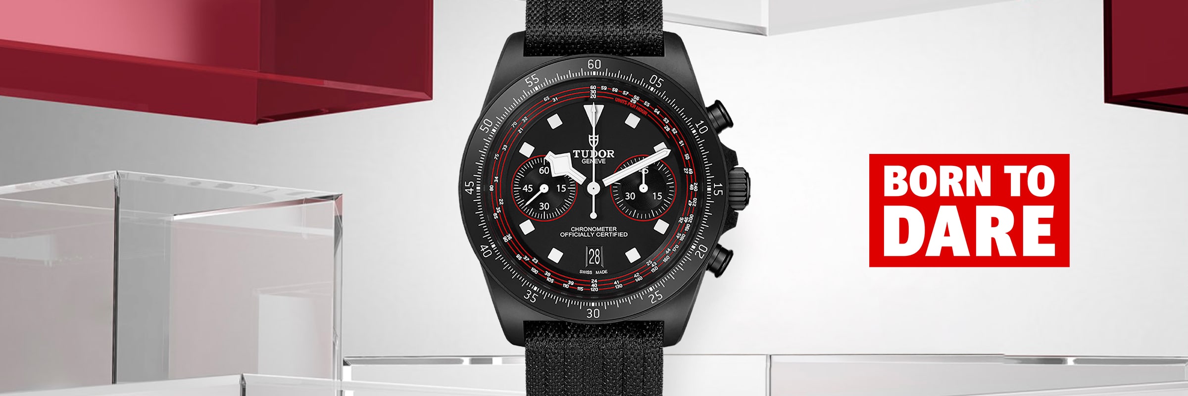 2025 © TUDOR Eine schwarze Tudor Pelagos FXD Chronographenuhr mit Stoffarmband steht aufrecht vor einem modernen, geometrischen Hintergrund, mit dem Slogan "BORN TO DARE" in fetter roter und weißer Schrift auf der rechten Seite.