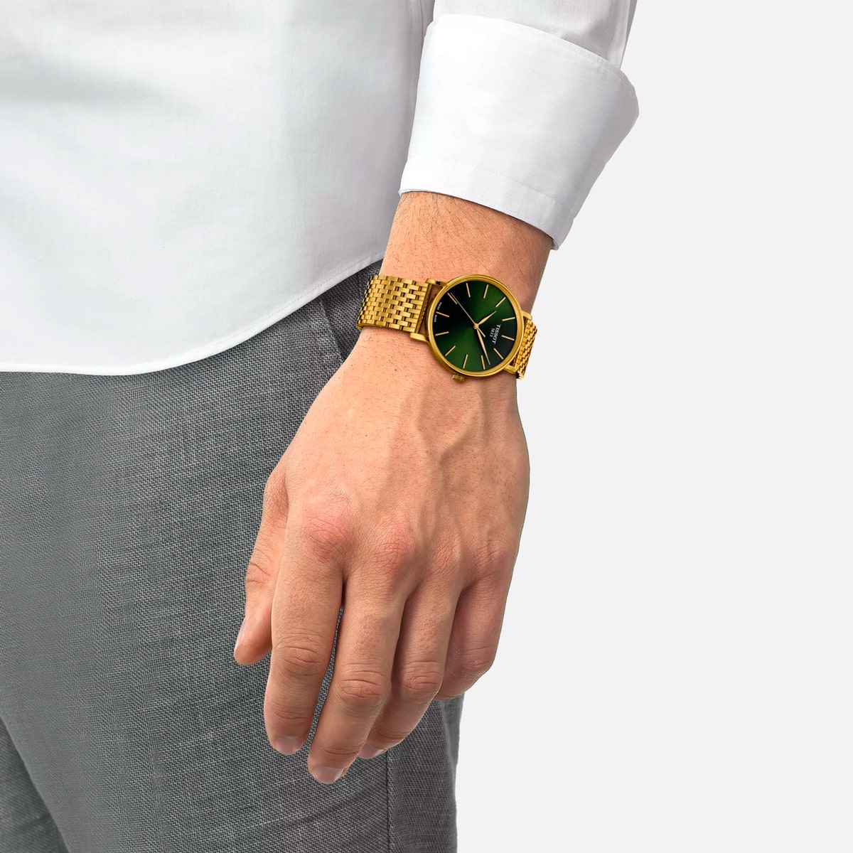2025 © TISSOT Eine Person, die ein weißes Hemd und eine graue Hose trägt, zeigt eine goldene Tissot Everytime-Armbanduhr mit grünem Zifferblatt und goldenen Details an ihrem linken Handgelenk. Die Hand ist entspannt und hängt an der Seite vor einem schlichten Hintergrund.