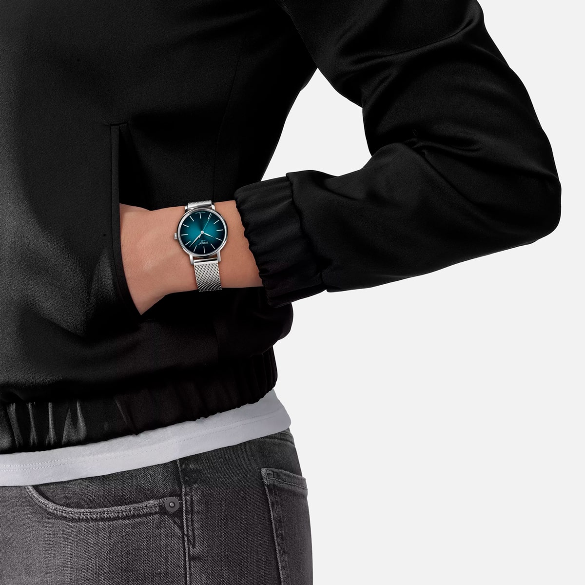 2025 © TISSOT Eine Person, die eine schwarze Jacke und eine graue Hose trägt, hat die linke Hand in der Tasche und zeigt eine silberne Tissot Everytime-Armbanduhr mit blauem Zifferblatt und Netzband.