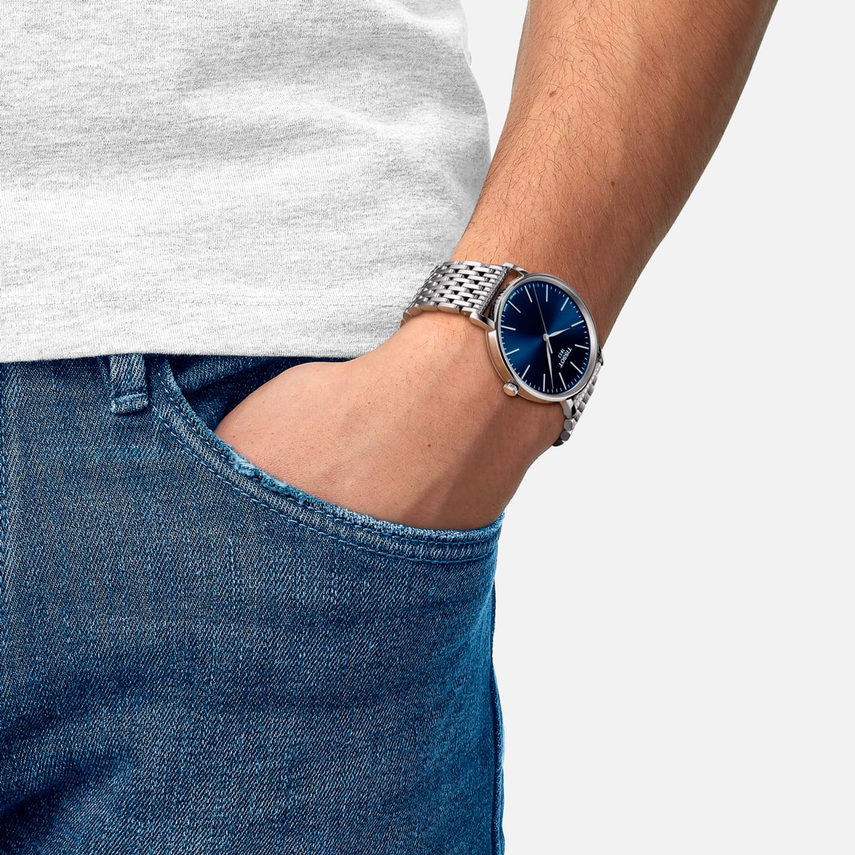 2025 © TISSOT Eine Person, die blaue Jeans und ein hellgraues T-Shirt trägt, hat die linke Hand in der Tasche und zeigt eine silberne Tissot Everytime-Armbanduhr mit blauem Zifferblatt und Metallarmband.