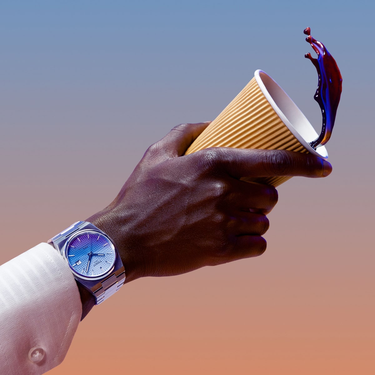 2025 © TISSOT Eine Hand, die eine silberne Tissot PRX-Armbanduhr trägt, kippt einen gelben Pappbecher um, woraufhin ein Spritzer dunkler Flüssigkeit austritt, vor einem Hintergrund mit Farbverlauf in Blau und Pfirsich.