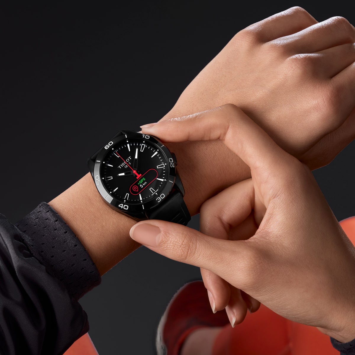 2025 © TISSOT Eine Person in einem orangefarbenen Outfit prüft die Zeit auf einer schwarzen Tissot T-Touch Armbanduhr mit roten Akzenten und stellt die Uhr mit der anderen Hand vor einem dunklen Hintergrund ein.