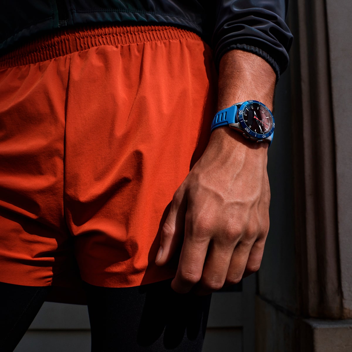 2025 © TISSOT Nahaufnahme der Hand und des Oberkörpers einer Person, die leuchtend orangefarbene Sporthosen, eine dunkle Jacke und eine blaue Tissot T-Touch-Armbanduhr trägt und im Freien in der Nähe eines Gebäudes steht.