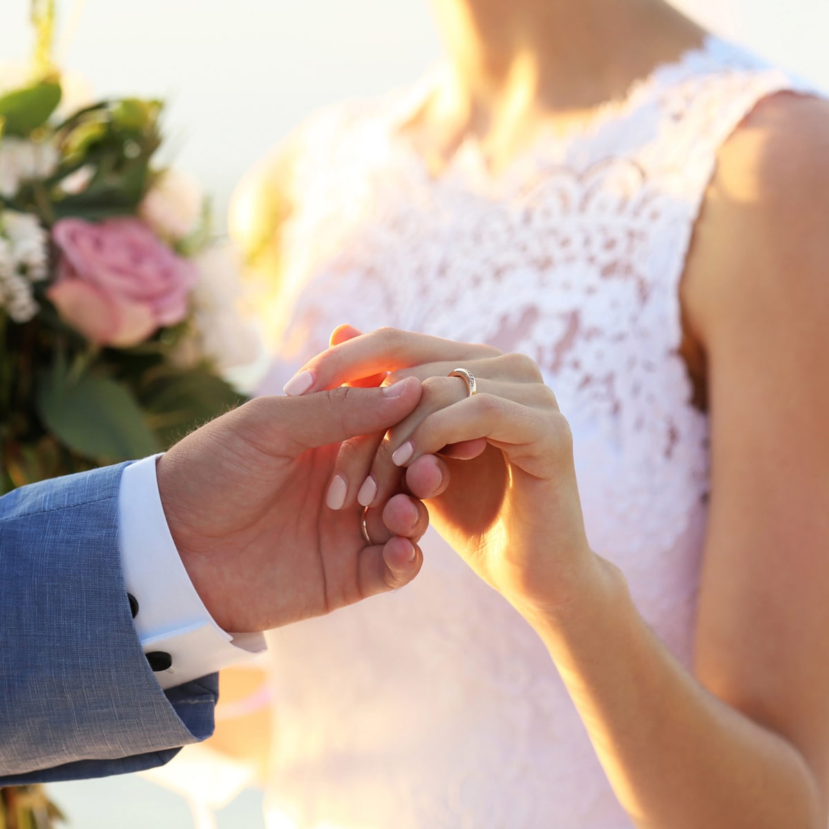 2025 © 123GOLD Eine Braut in einem weißen Spitzenkleid hält einen Blumenstrauß und berührt sanft die Hände des Bräutigams in einem blauen Anzug während ihrer Trauung, wobei sie ihren Ehering zeigt.