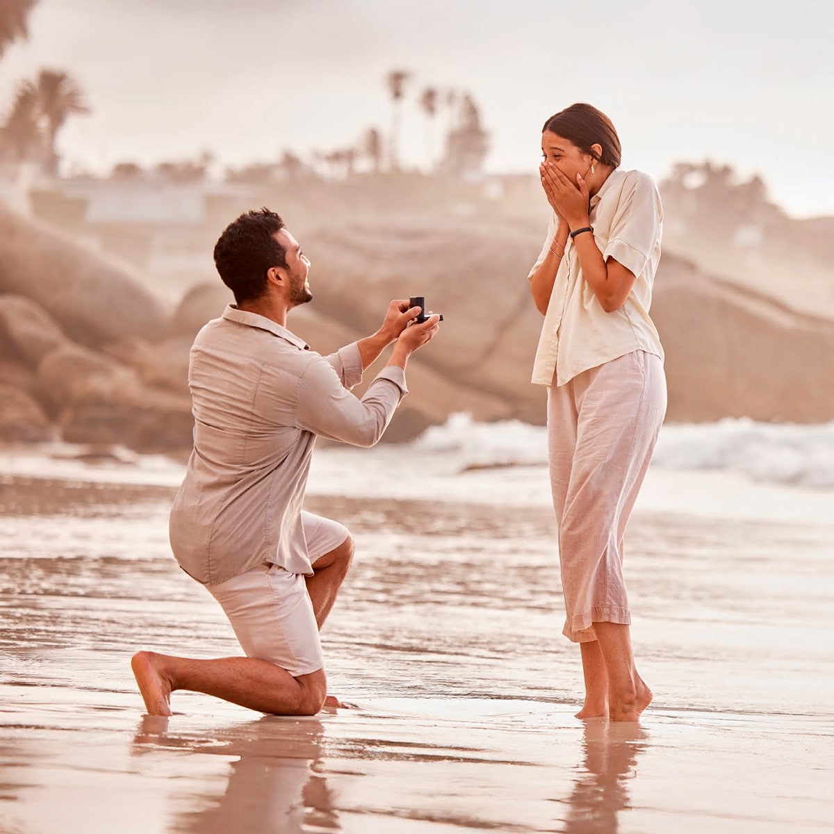 2025 © 123GOLD Ein Mann kniet am Strand mit einer offenen Ringschachtel in der Hand und macht einer überraschten Frau, die ihren Mund mit den Händen bedeckt, einen Antrag. Im Hintergrund sind das Meer und die Felsen bei Sonnenuntergang zu sehen, die einen romantischen Moment vor ihrer Trauung festhalten.