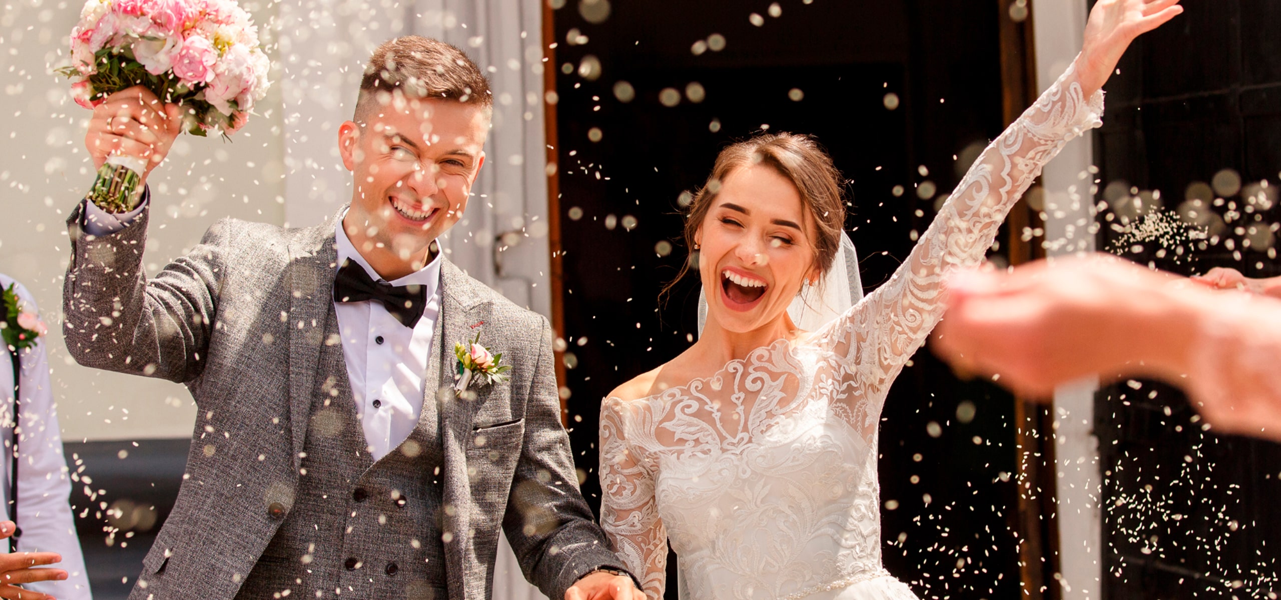 2025 © 123GOLD Ein fröhliches Brautpaar in Hochzeitskleidung feiert draußen nach der Trauung mit einem breiten Lächeln, während die Gäste sie mit Konfetti bewerfen. Die Braut hebt aufgeregt einen Arm und hält die Hand des Bräutigams.