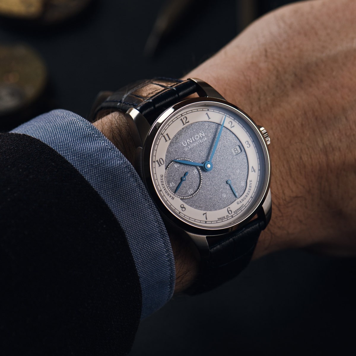 2025 © UNION GLASHÜTTE Eine Person, die ein formelles Hemd und ein Jackett trägt, zeigt eine Union Glashütte 1893 Armbanduhr mit blauem Armband und einem silber-blauen Zifferblatt mit drei Hilfszifferblättern und blauen Zeigern.