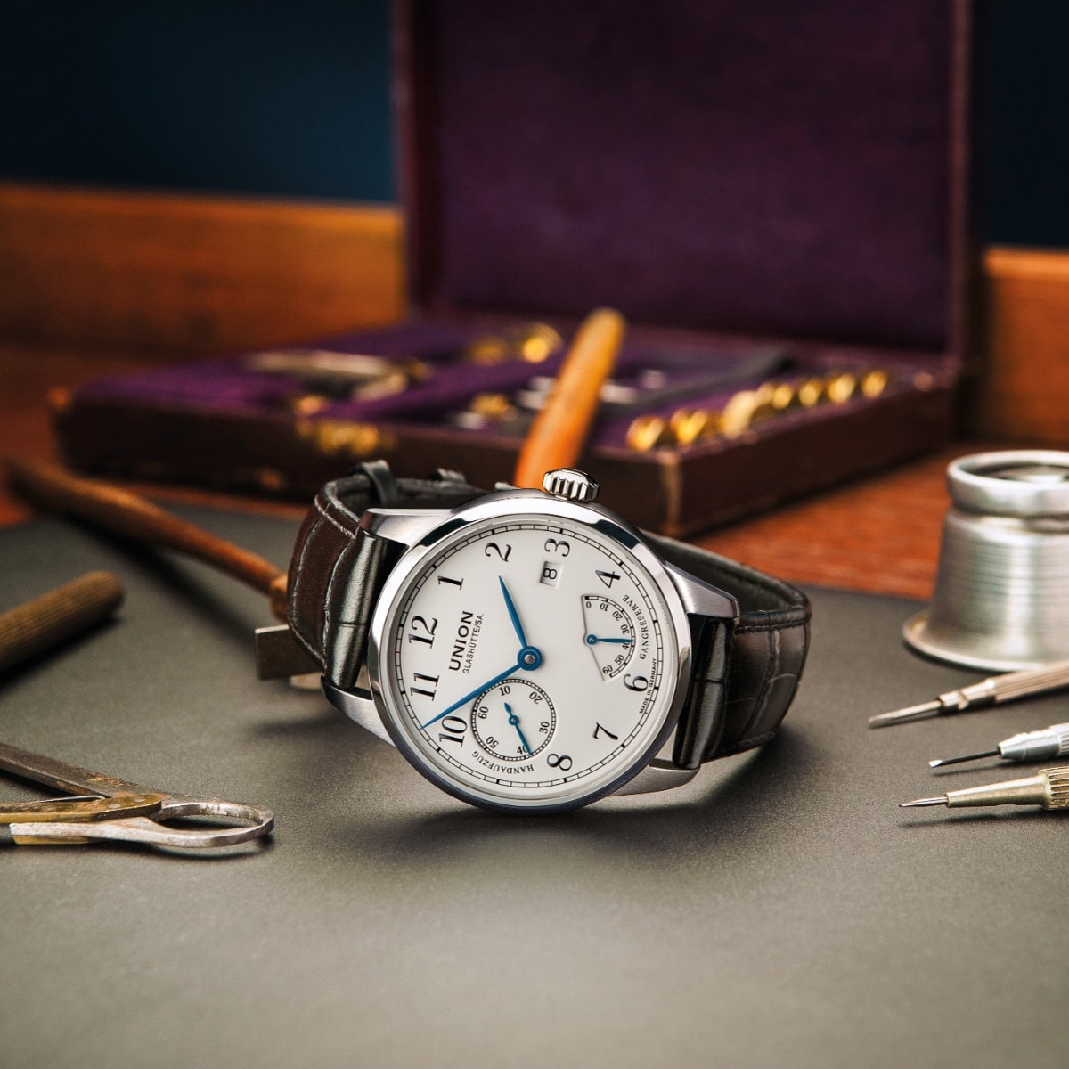 2025 © UNION GLASHÜTTE Eine luxuriöse Union Glashütte 1893-Armbanduhr mit weißem Zifferblatt und braunem Lederarmband liegt auf einem Schreibtisch, umgeben von Uhrmacherwerkzeugen, Garn und einer Holzkiste. Das Zifferblatt ist mit arabischen Ziffern und zwei Hilfszifferblättern versehen.