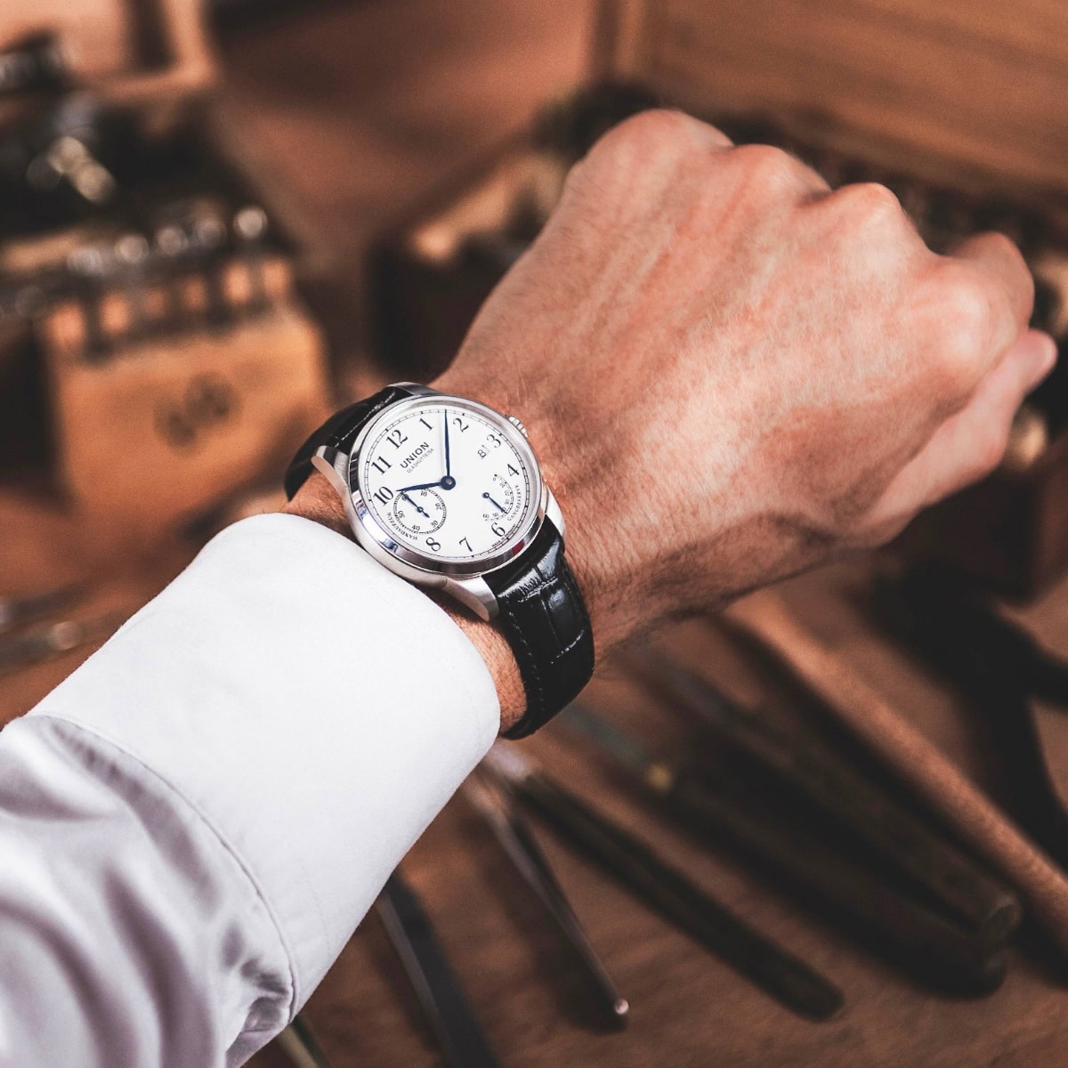 2025 © UNION GLASHÜTTE Eine Person, die ein weißes Hemd mit Manschetten trägt, zeigt eine klassische Union Glashütte 1893 Armbanduhr mit schwarzem Lederarmband und weißem Zifferblatt, die die Zeit 10:09 anzeigt. Im Hintergrund sind unscharfe Werkzeuge und eine Holzkiste zu sehen.
