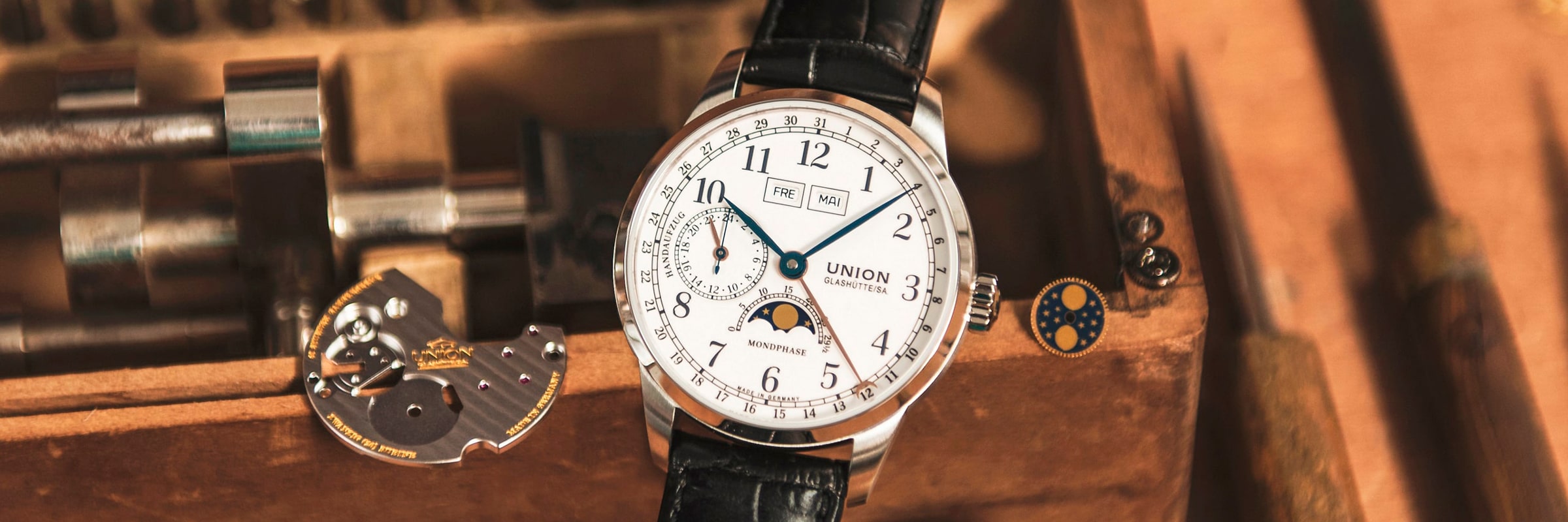 2025 © UNION GLASHÜTTE Nahaufnahme einer Luxusarmbanduhr Union Glashütte 1893 mit weißem Zifferblatt, schwarzen Ziffern und mehreren Zifferblättern, die auf einer hölzernen Unterlage neben Uhrmacherwerkzeugen und kleinen mechanischen Teilen liegt.