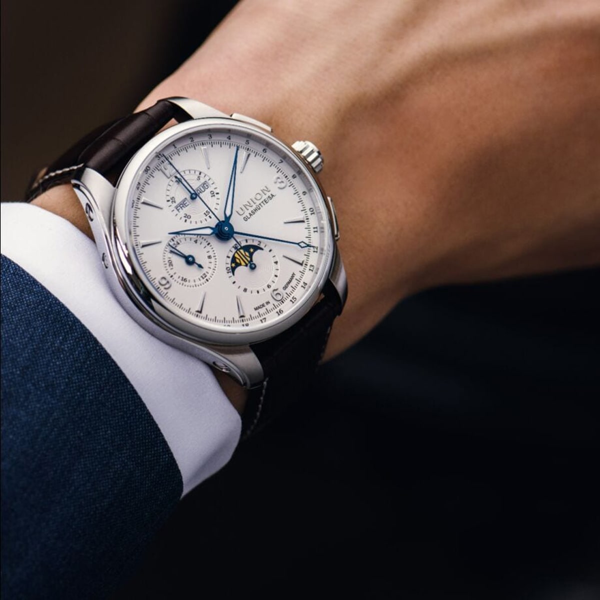 2025 © UNION GLASHÜTTE Nahaufnahme des Handgelenks einer Person, die eine silber-weiße Analoguhr Union Glashütte Belisar mit Mondphasenanzeige, mehreren Hilfszifferblättern, Lederarmband und formellem blauen Anzugärmel trägt.