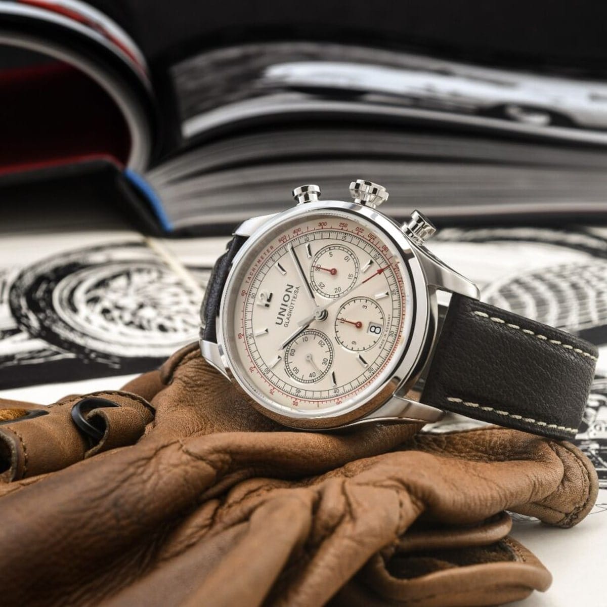 2025 © UNION GLASHÜTTE Eine silberne Union Glashütte Belisar Armbanduhr mit weißem Zifferblatt und Chronographenfunktion ruht auf braunen Lederhandschuhen, im Hintergrund ein aufgeschlagenes Buch mit schwarz-weißen Illustrationen.