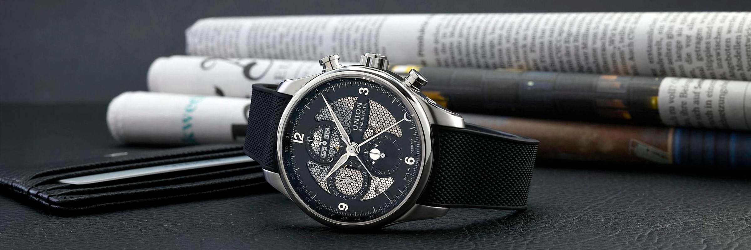 2025 © UNION GLASHÜTTE Eine schwarz-silberne Union Glashütte Belisar-Armbanduhr mit strukturiertem Zifferblatt und Chronographenzifferblättern liegt auf einer dunklen Fläche neben einer Lederbörse, im Hintergrund liegen aufgerollte Zeitungen.