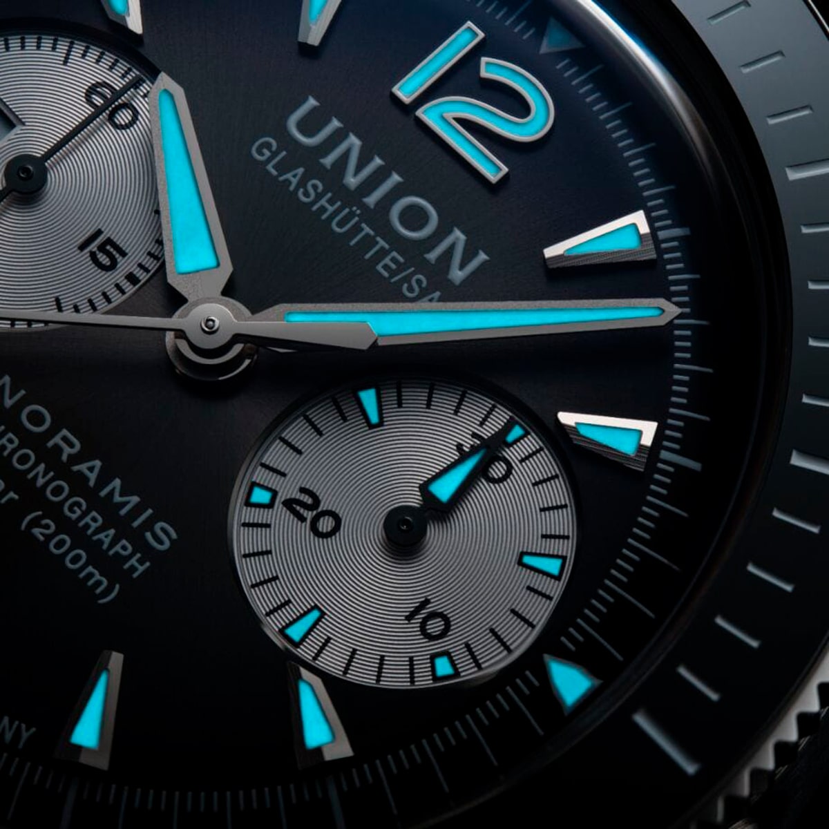 2025 © UNION GLASHÜTTE Nahaufnahme des Zifferblatts einer Union Glashütte Noramis mit schwarzem Zifferblatt, blauen Leuchtmarkierungen, Hilfszifferblättern und silbernen Zeigern, mit der Nummer 12 an der Spitze.