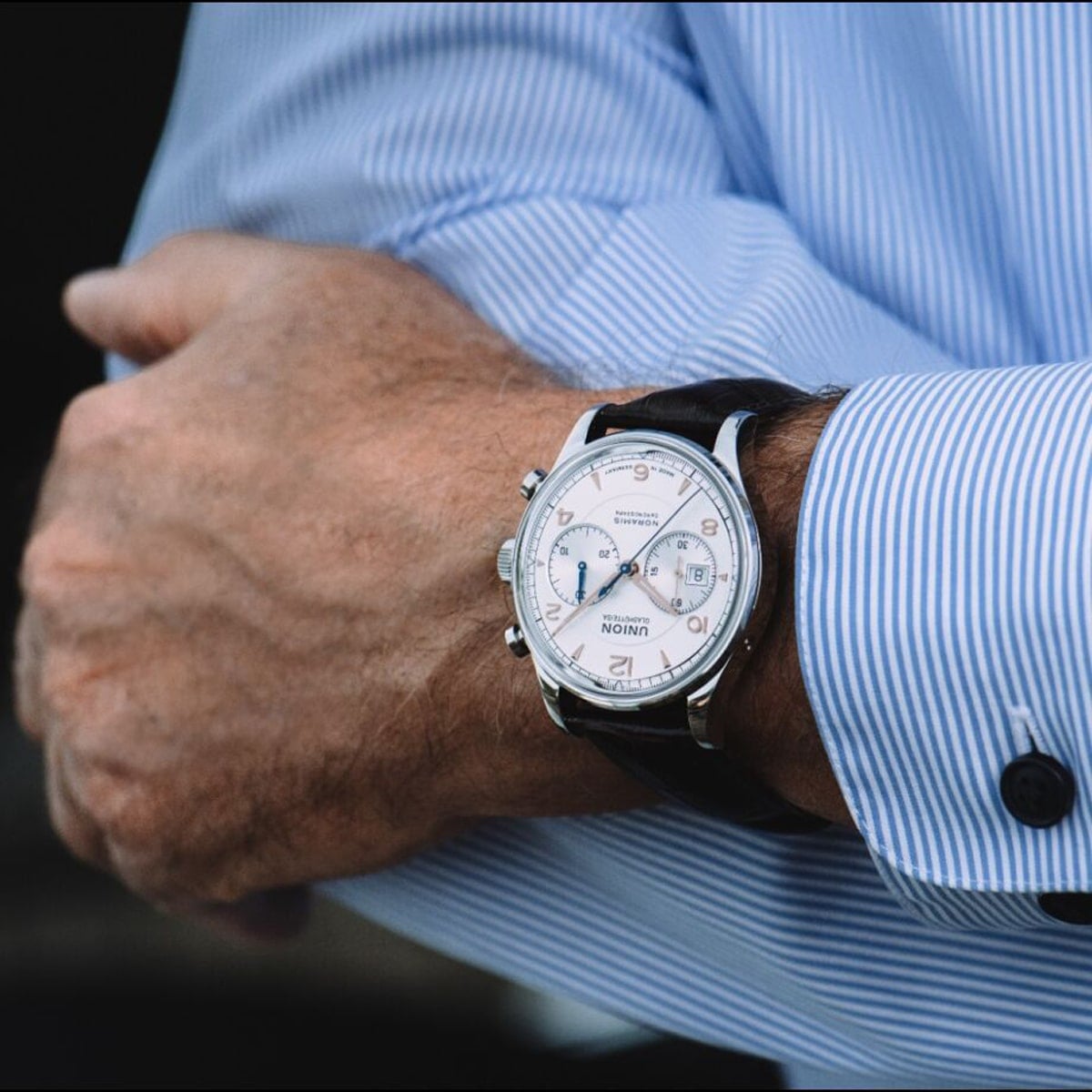2025 © UNION GLASHÜTTE Eine Person, die ein blaues Nadelstreifenhemd trägt, rückt ihren Ärmel zurecht und enthüllt eine silberne Union Glashütte Noramis Armbanduhr mit weißem Zifferblatt und schwarzem Lederarmband.
