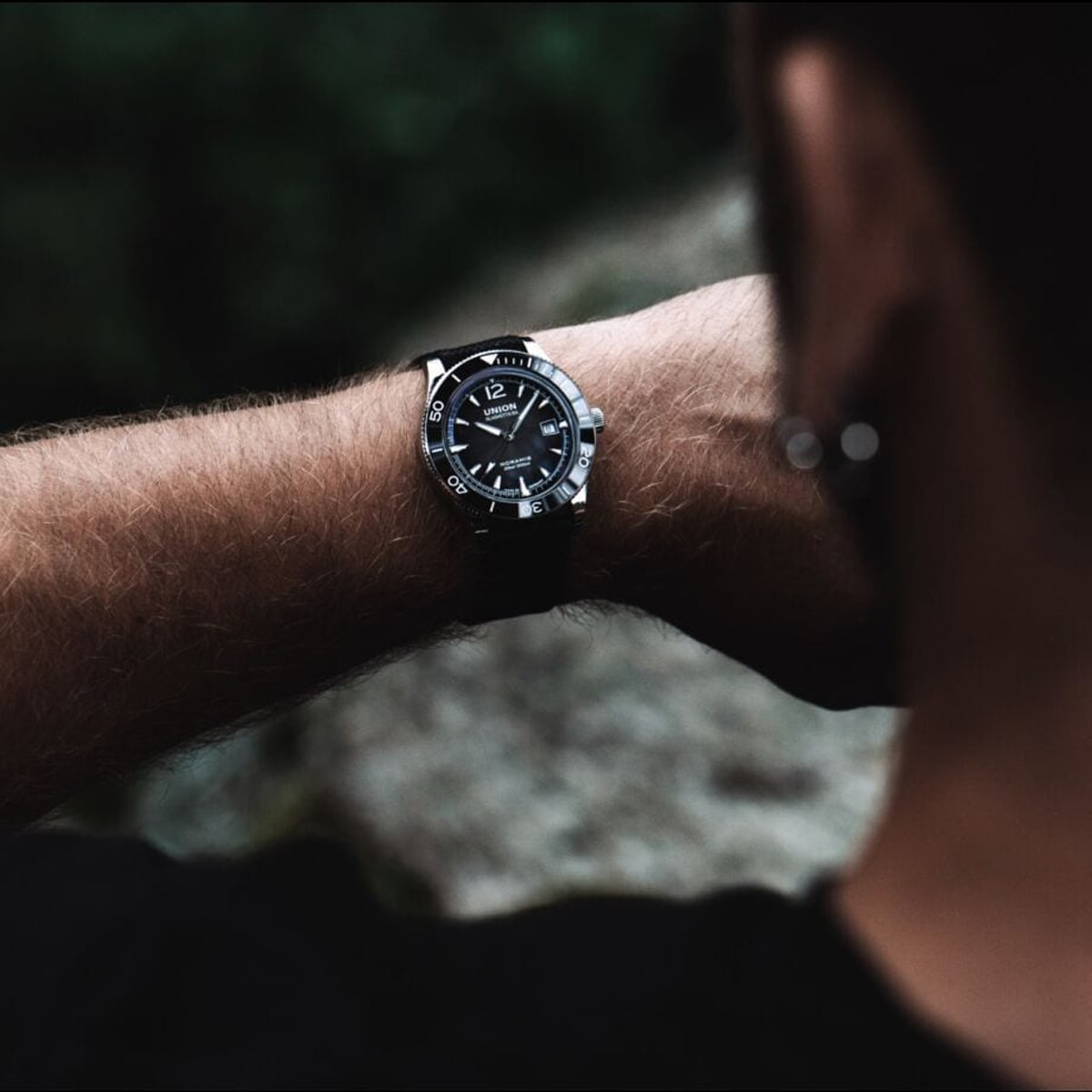 2025 © UNION GLASHÜTTE Eine Person mit Bart und Ohrring blickt auf eine schwarze Union Glashütte Noramis-Armbanduhr an ihrem linken Handgelenk, im Freien mit Grün im unscharfen Hintergrund.