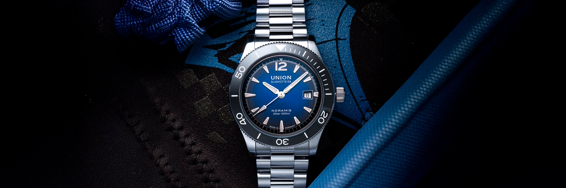 2025 © UNION GLASHÜTTE Eine Union Glashütte Noramis Edelstahl-Armbanduhr mit blauem Zifferblatt und Lünette, silbernen Indexen und einem Metallarmband, dargestellt auf einem dunklen Hintergrund mit blauen und schwarzen Stoffakzenten.