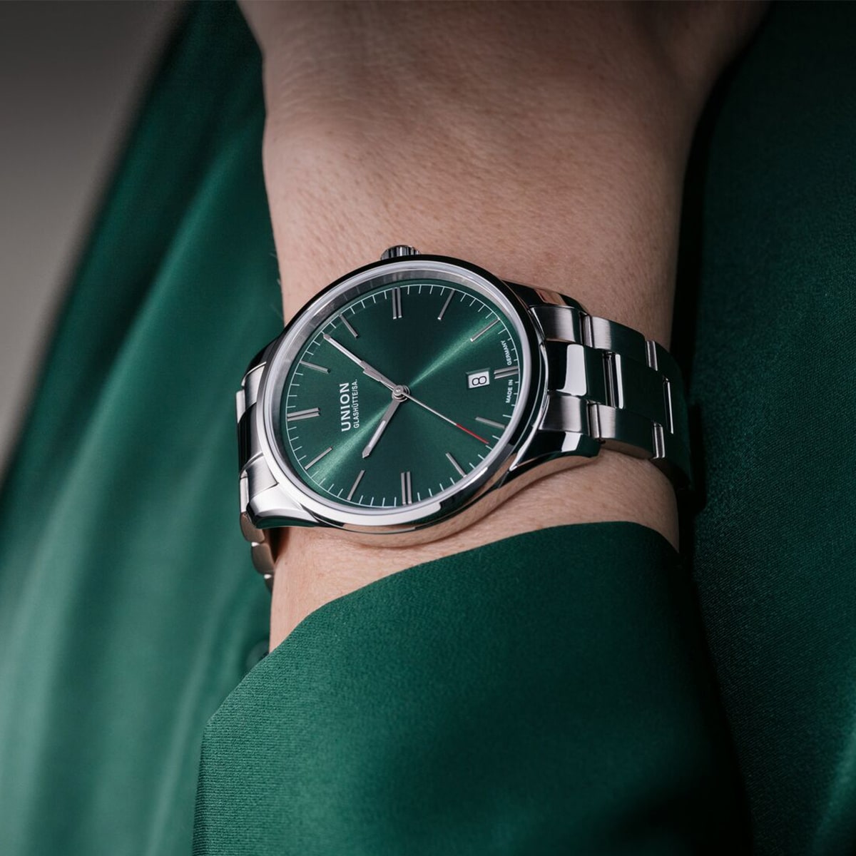 2025 © UNION GLASHÜTTE Nahaufnahme einer Person, die eine silberne Union Glashütte Viro-Armbanduhr mit grünem Zifferblatt und Datumsanzeige trägt, ergänzt durch ein grünes Outfit, das das elegante und moderne Design der Uhr hervorhebt.