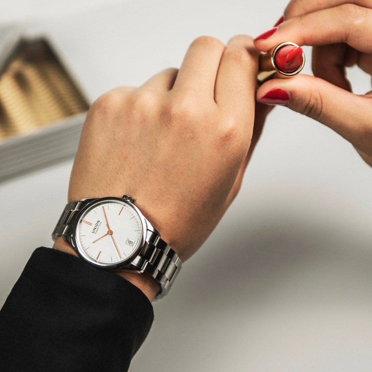 2025 © UNION GLASHÜTTE Eine Person mit rot lackierten Nägeln bereitet sich darauf vor, roten Lippenstift aufzutragen. Sie trägt eine silberne Armbanduhr von Union Glashütte Viro und einen dunklen Ärmel, im Hintergrund ist eine verschwommene Schachtel zu sehen.