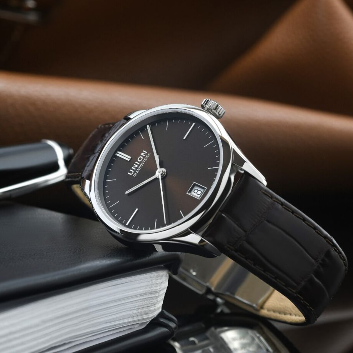 2025 © UNION GLASHÜTTE Eine luxuriöse Union Glashütte Viro Armbanduhr mit schwarzem Lederarmband und schwarzem Zifferblatt ruht auf einem geschlossenen Notizbuch, wobei ein Stift und braune Ledergegenstände im Hintergrund verschwimmen. Die Uhr zeigt das Datum auf der 3-Uhr-Position an.