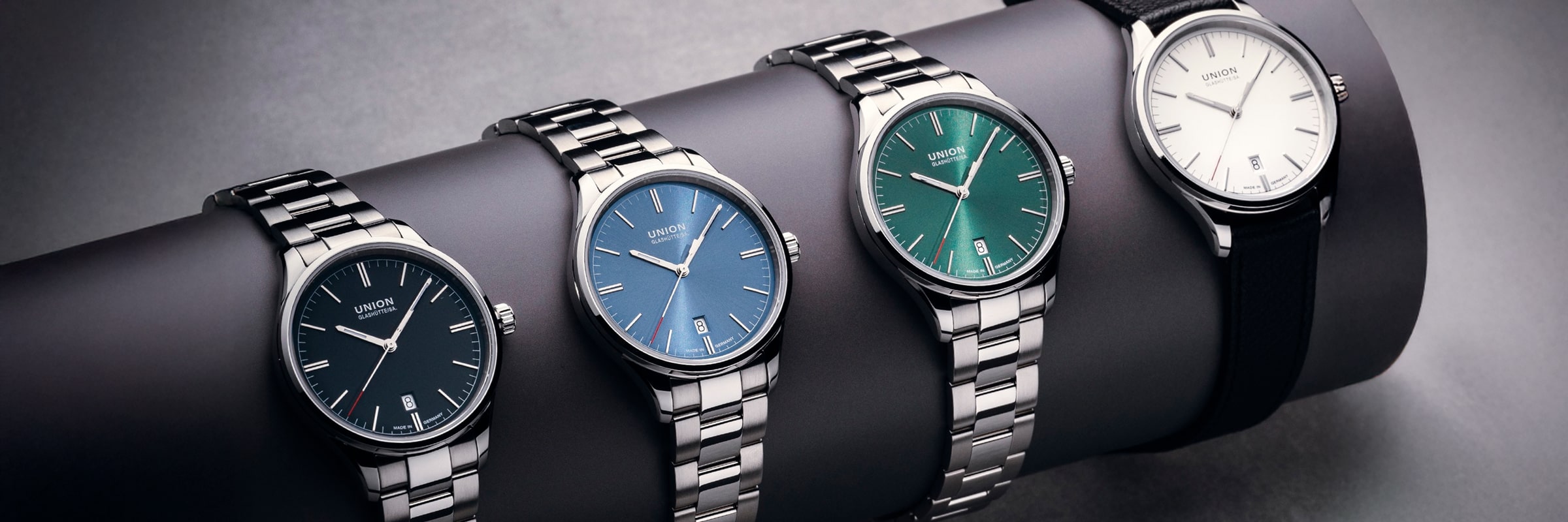 2025 © UNION GLASHÜTTE Vier Union Glashütte Viro Armbanduhren mit silbernen Armbändern und farbigen Zifferblättern - schwarz, blau, grün und weiß - werden auf einem zylindrischen Ständer präsentiert. Die Uhr mit dem weißen Zifferblatt hat ein klassisches schwarzes Lederarmband anstelle von Metall.