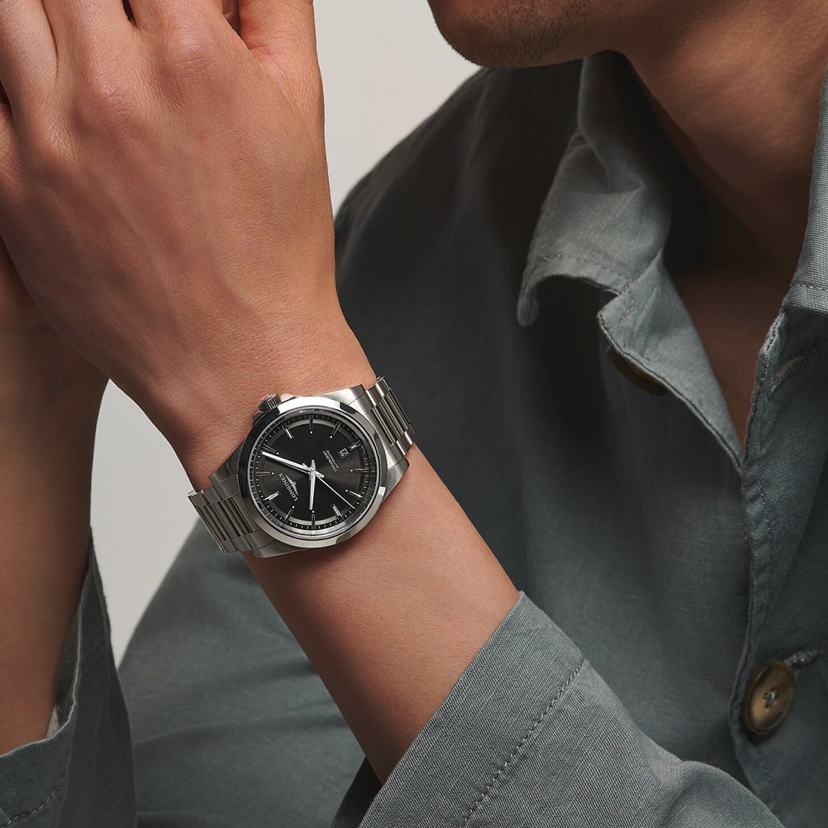 2025 © LONGINES Eine Person, die eine silberne LONGINES-Armbanduhr mit schwarzem Zifferblatt trägt, bekleidet mit einem hellgrauen Button-up-Hemd. Die Hände der Person sind in der Nähe ihres Gesichts erhoben, wodurch die Uhr an ihrem linken Handgelenk hervorgehoben wird.