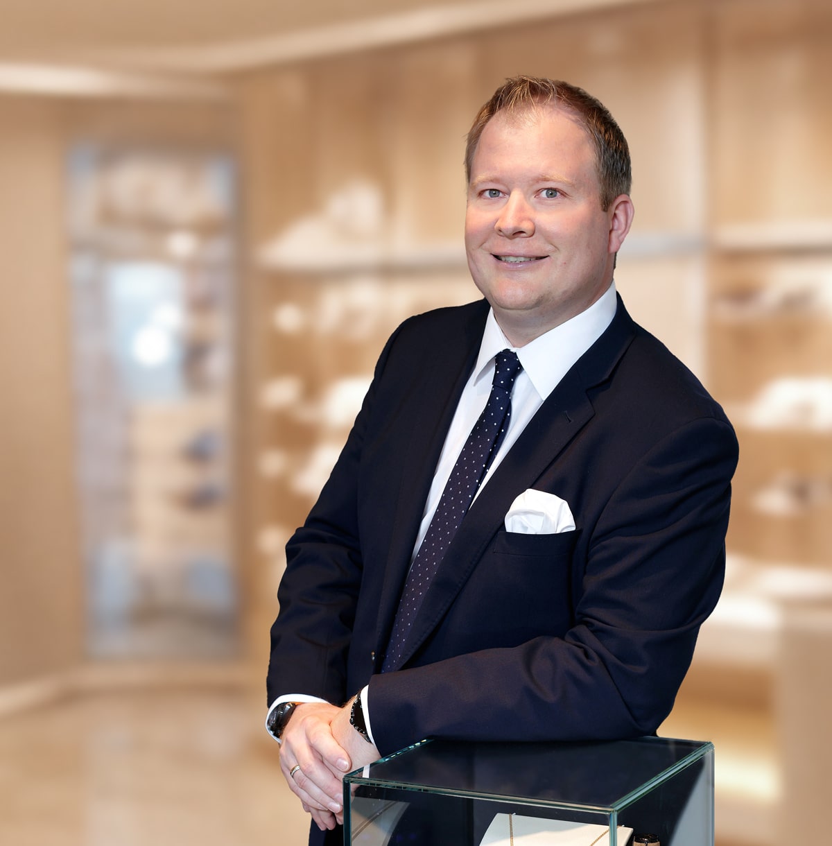 © Juwelier Wenthe Ein Mann in einem dunklen Anzug, weißem Hemd und Krawatte steht lächelnd an eine Glasvitrine gelehnt in einem modernen, gut beleuchteten Raum mit verschwommenen Regalen im Hintergrund.