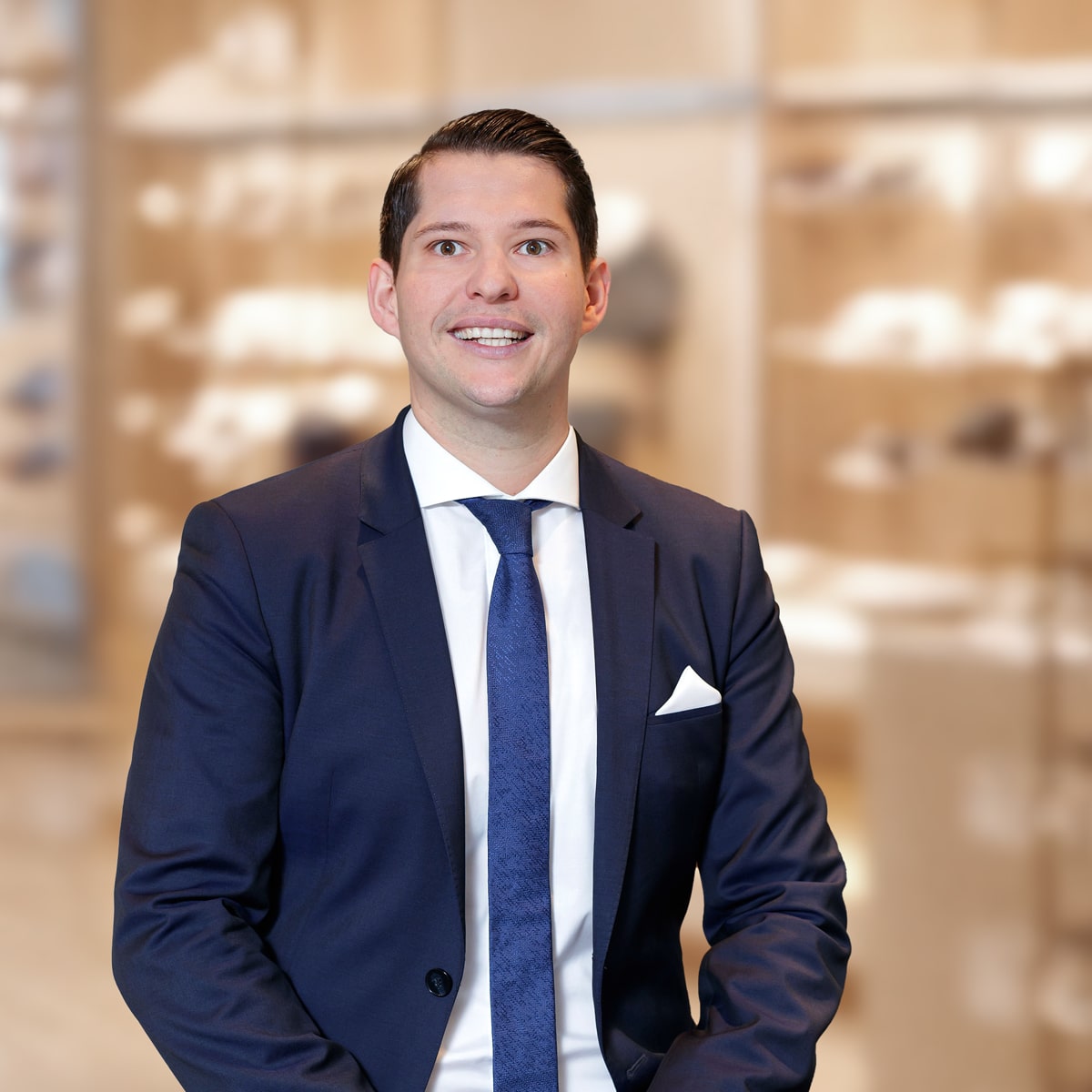© Juwelier Wenthe Ein lächelnder Mann in einem dunkelblauen Anzug, weißem Hemd und blauer Krawatte steht vor einem sanft verschwommenen Hintergrund mit Regalen und warmer Beleuchtung.