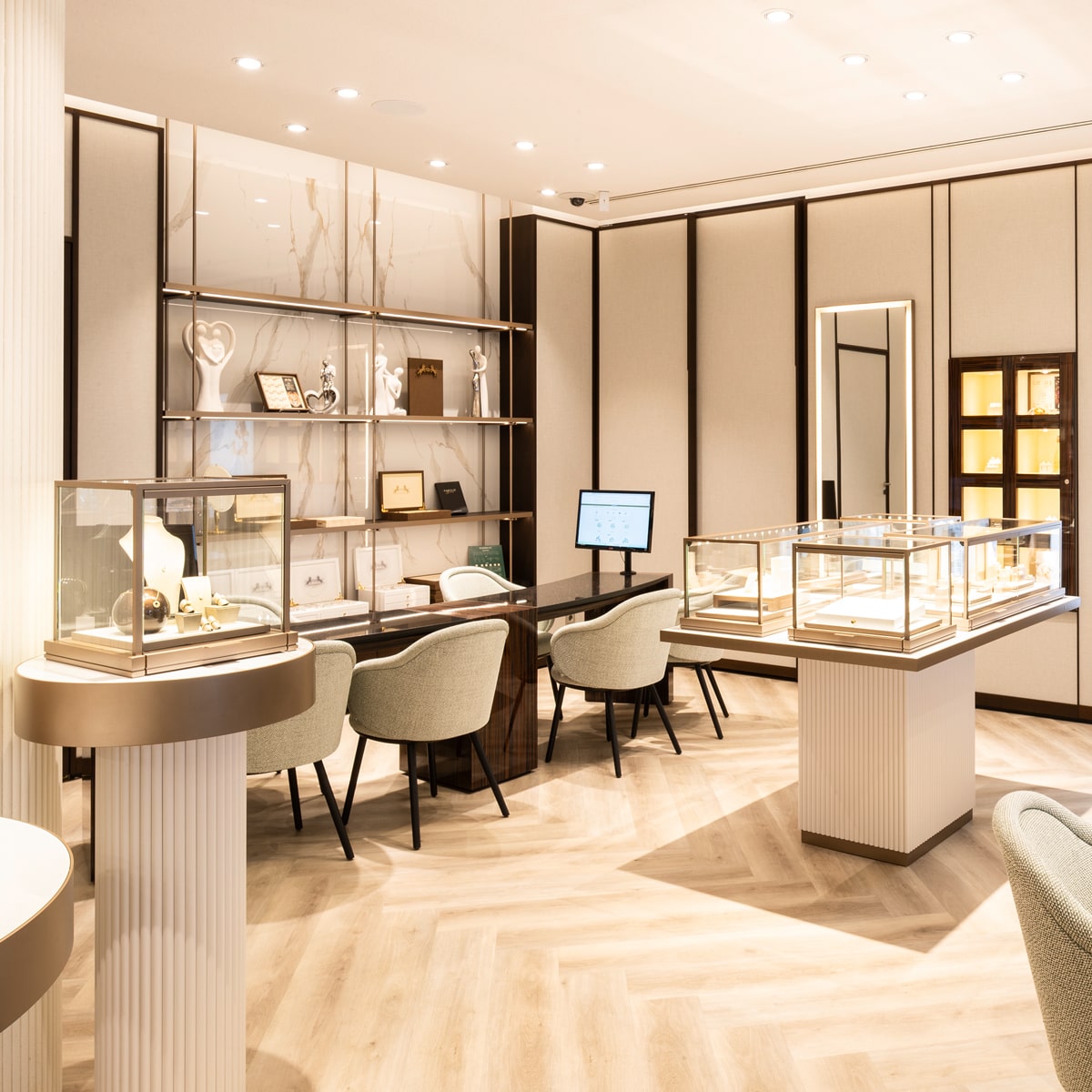 © Juwelier Wenthe Ein modernes, elegantes Juweliergeschäft mit Glasvitrinen, in denen hochwertige Schmuckmarken ausgestellt sind, beigen Stühlen, Holzregalen mit Dekorationsartikeln, einem Computer auf dem Schreibtisch und warmer Umgebungsbeleuchtung.
