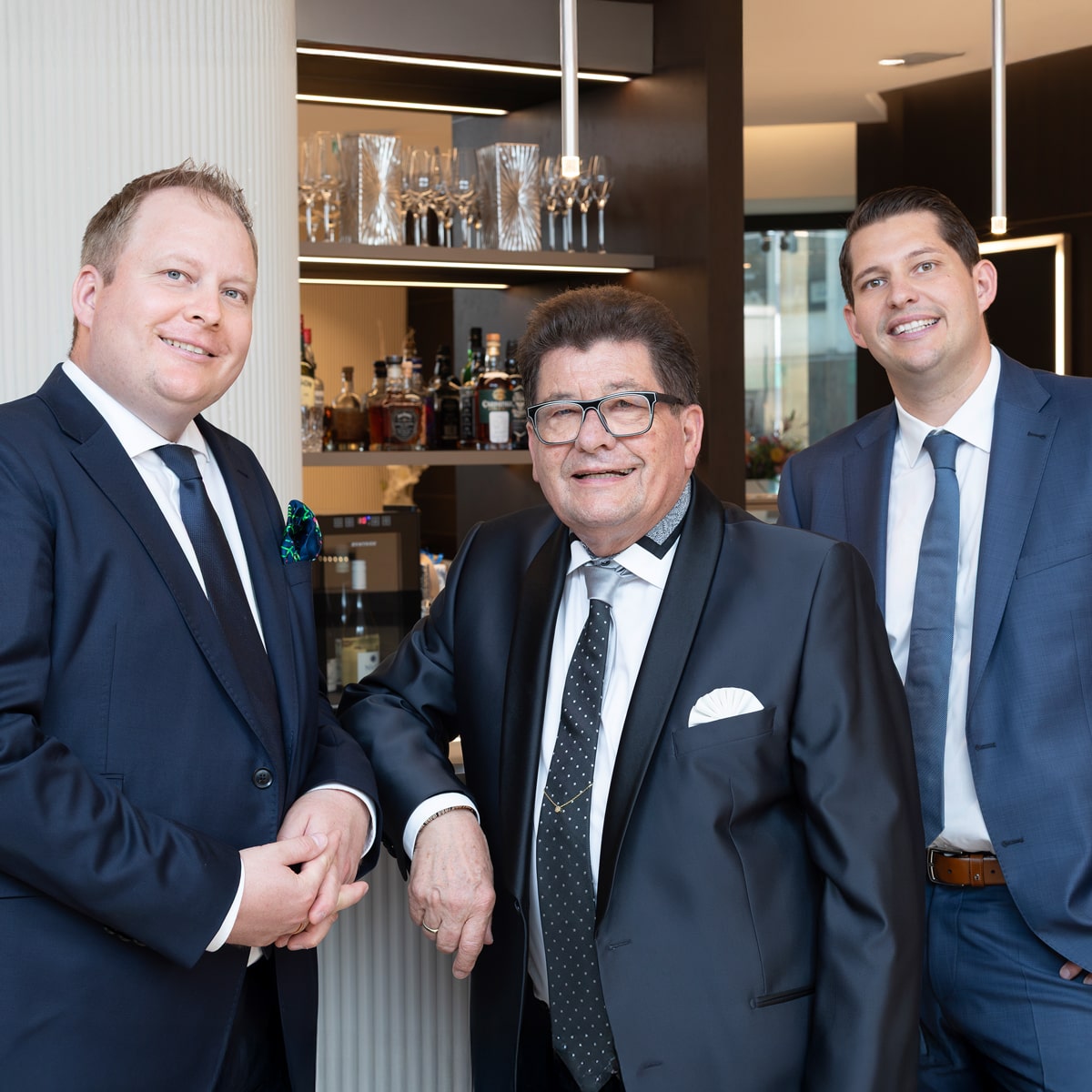 © Juwelier Wenthe Drei Männer in Anzügen stehen zusammen und lächeln in einer modernen Bar oder Lounge; hinter ihnen sind Gläser und Flaschen auf Regalen zu sehen.
